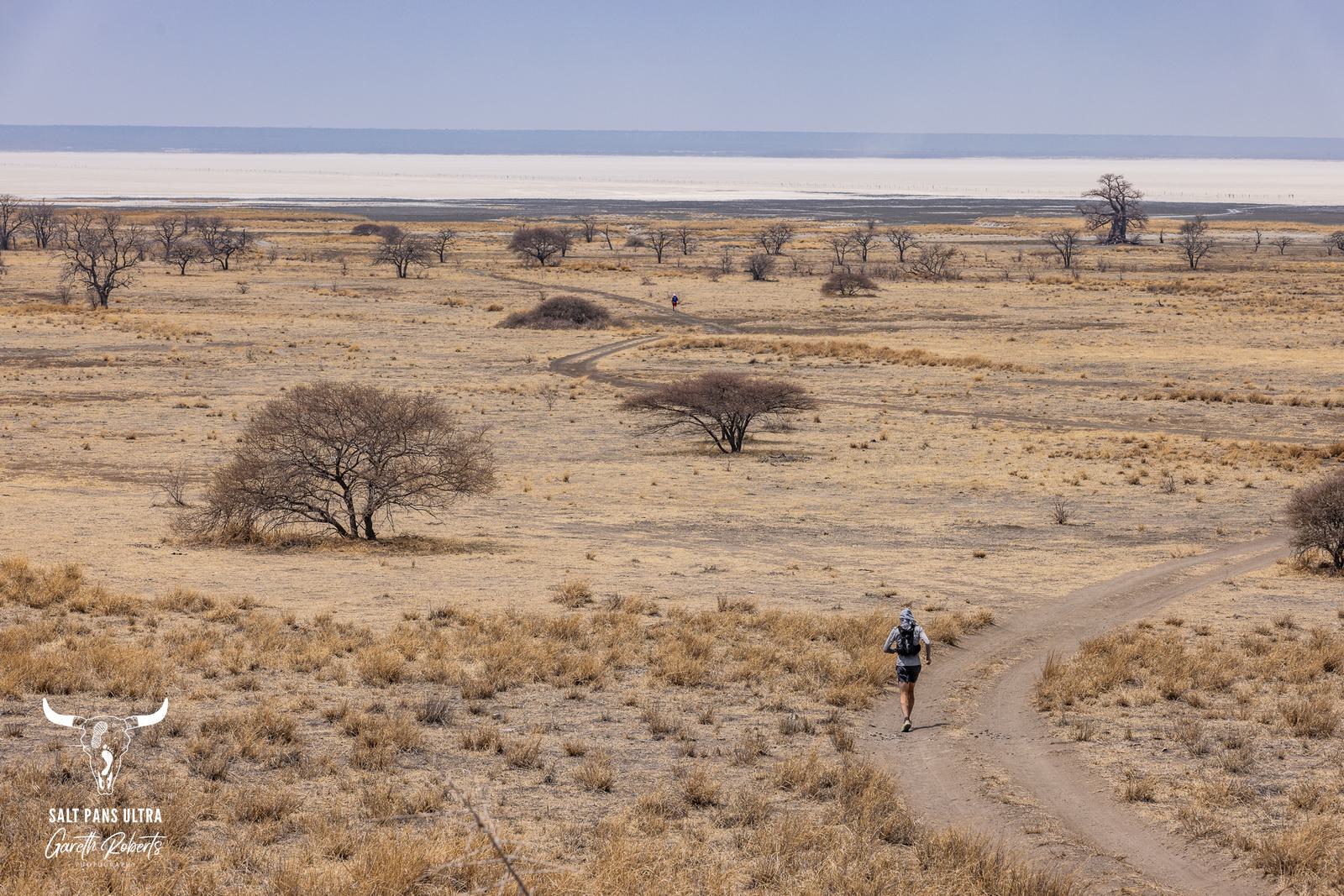 A person running on a dirt path in a vast, arid landscape with sparse trees and dry grass. The horizon features a large, flat expanse, possibly a salt pan, under a clear sky. A logo is in the bottom-left corner.