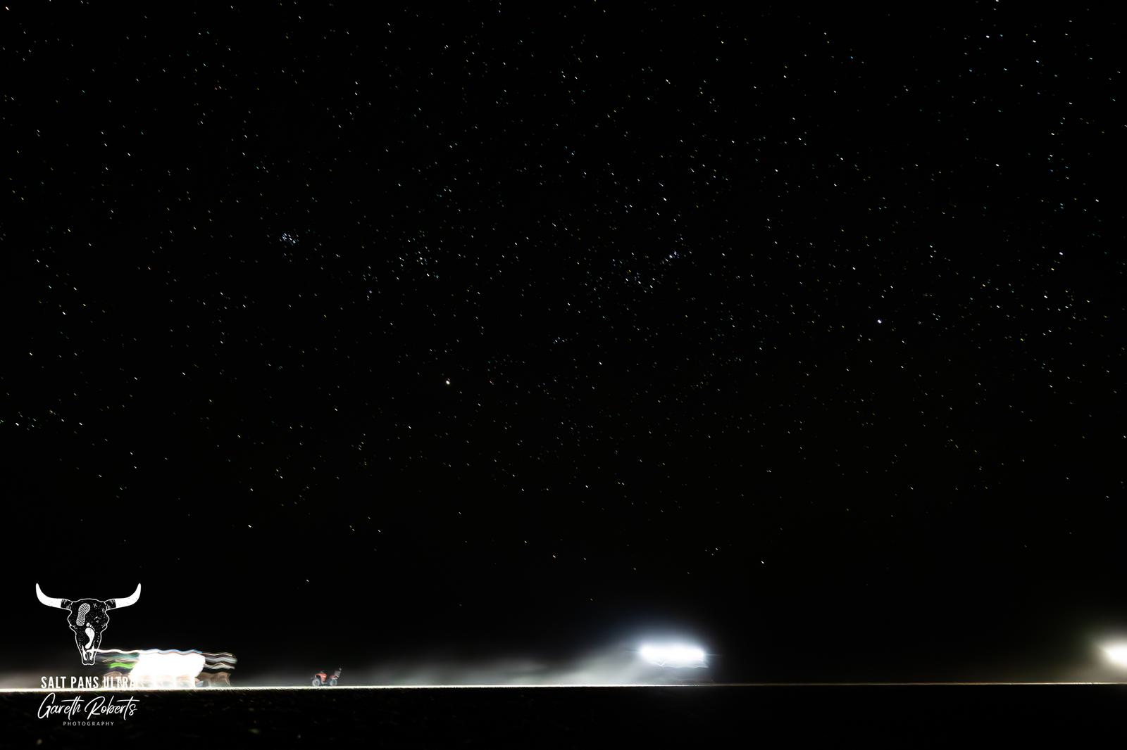 Starry night sky over a vast, flat landscape with distant bright lights near the horizon. The scene conveys a sense of calm and openness, with the silhouette of a bull logo in the bottom left corner.