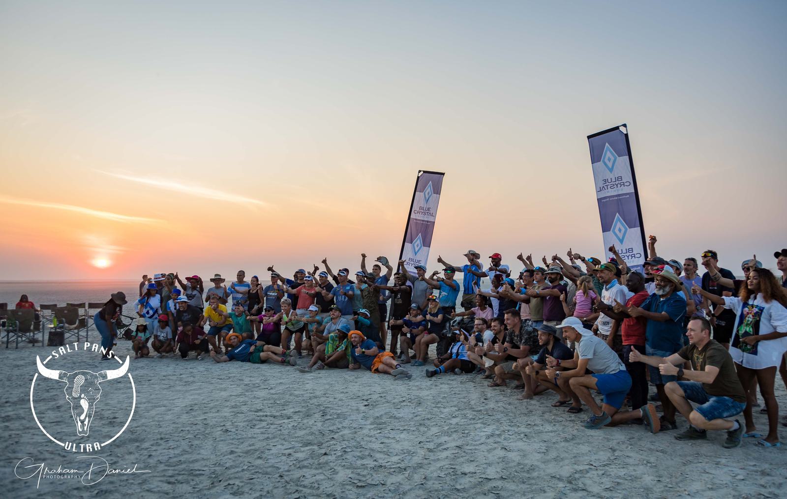 A large group of people gathered on a beach at sunset, celebrating and posing with two banners. They appear enthusiastic, some pointing or holding drinks. A logo with a bull and text is visible in the foreground.