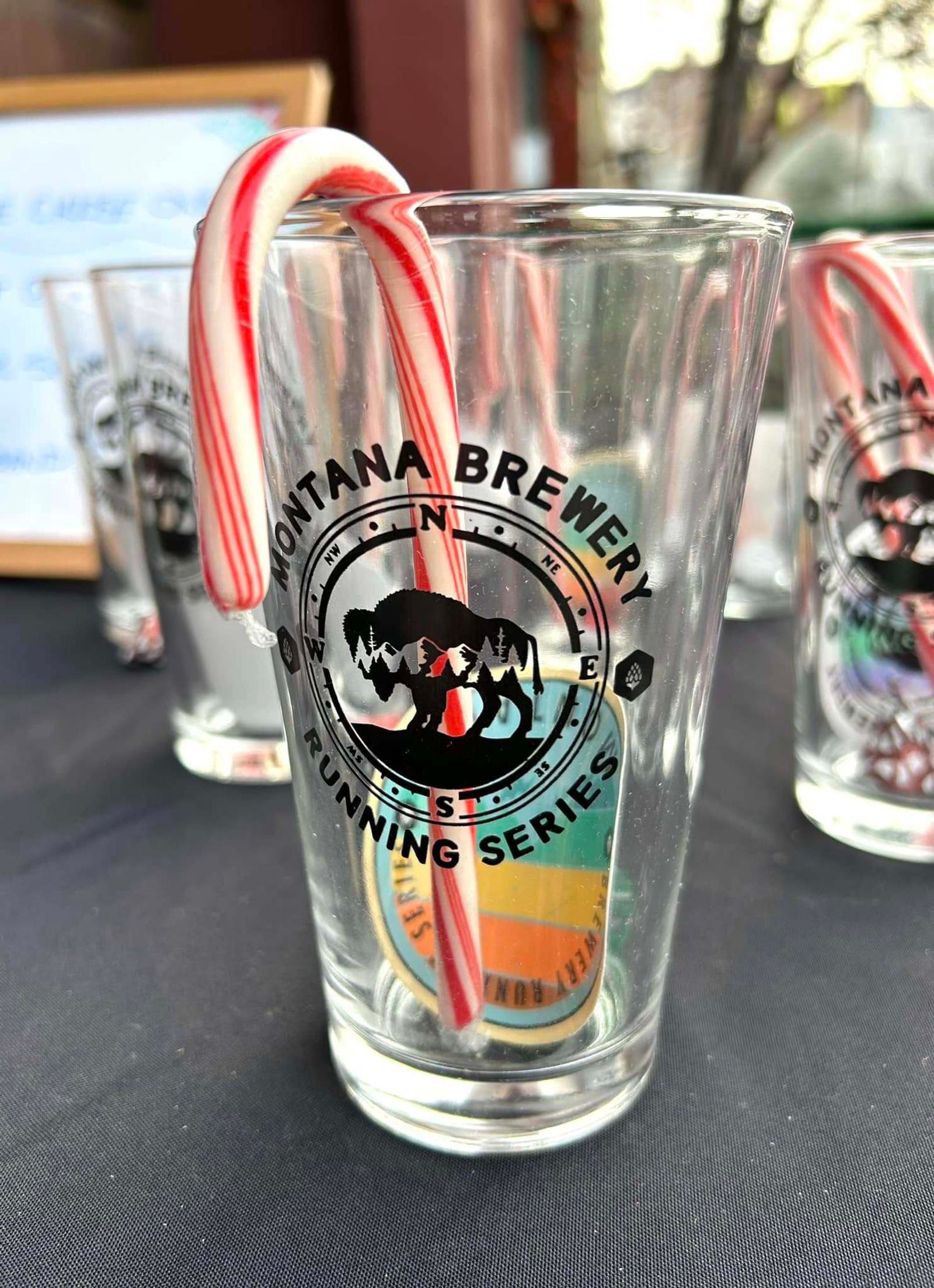 A glass with the Montana Brewery Running Series logo holds a candy cane. Other similar glasses are on a black tablecloth in the background.