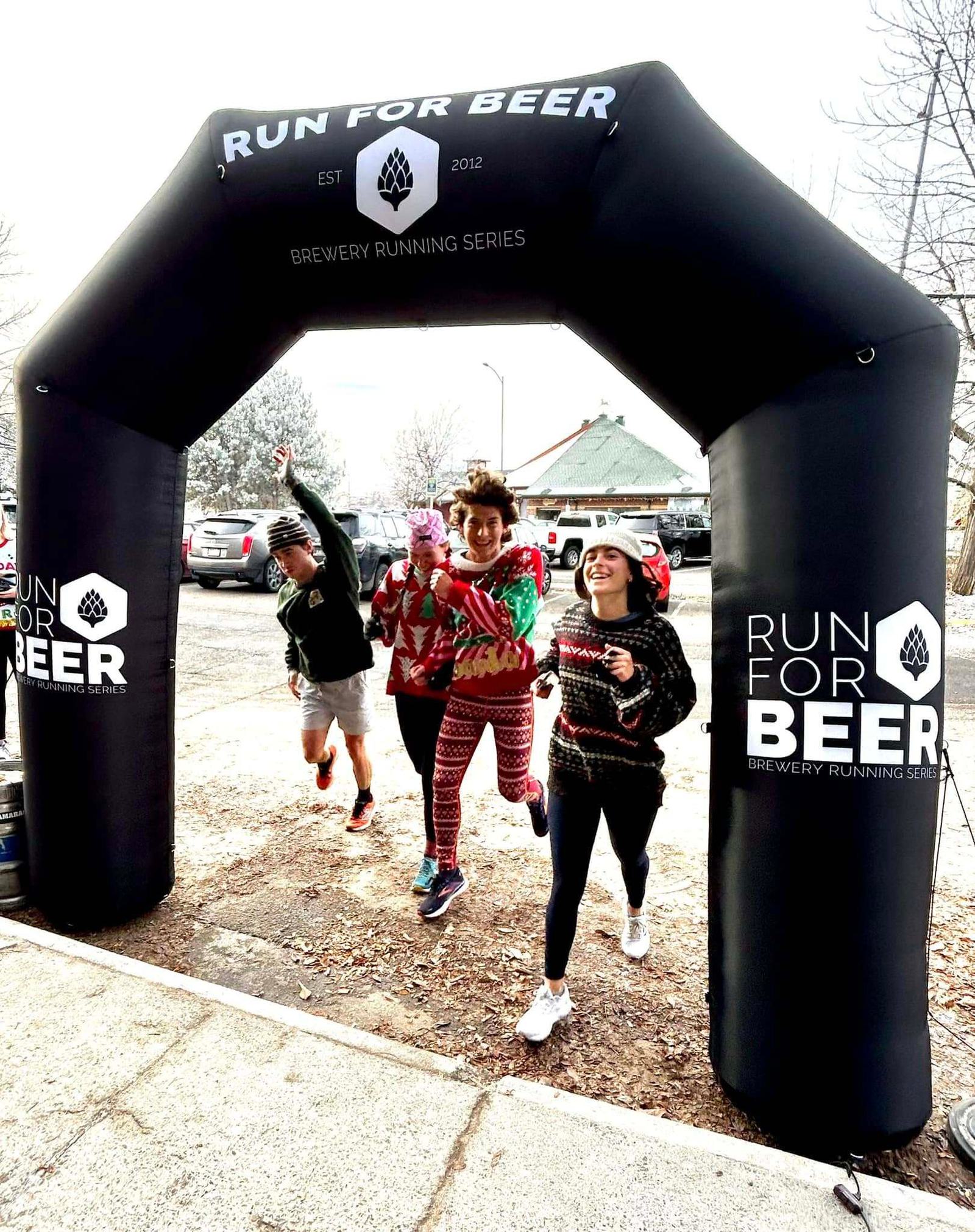 Four people wearing festive outfits joyfully run under a large black inflatable arch that reads "RUN FOR BEER - Brewery Running Series" at an outdoor event.