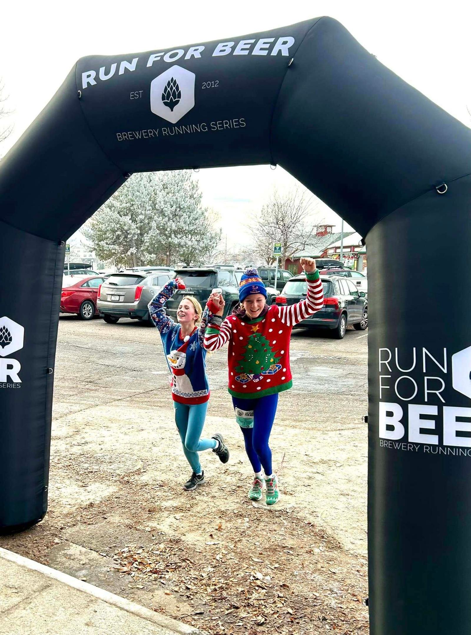 Two people wearing festive holiday sweaters and race bibs raise their arms in celebration as they cross the finish line under a black "Run for Beer" arch at an outdoor running event.