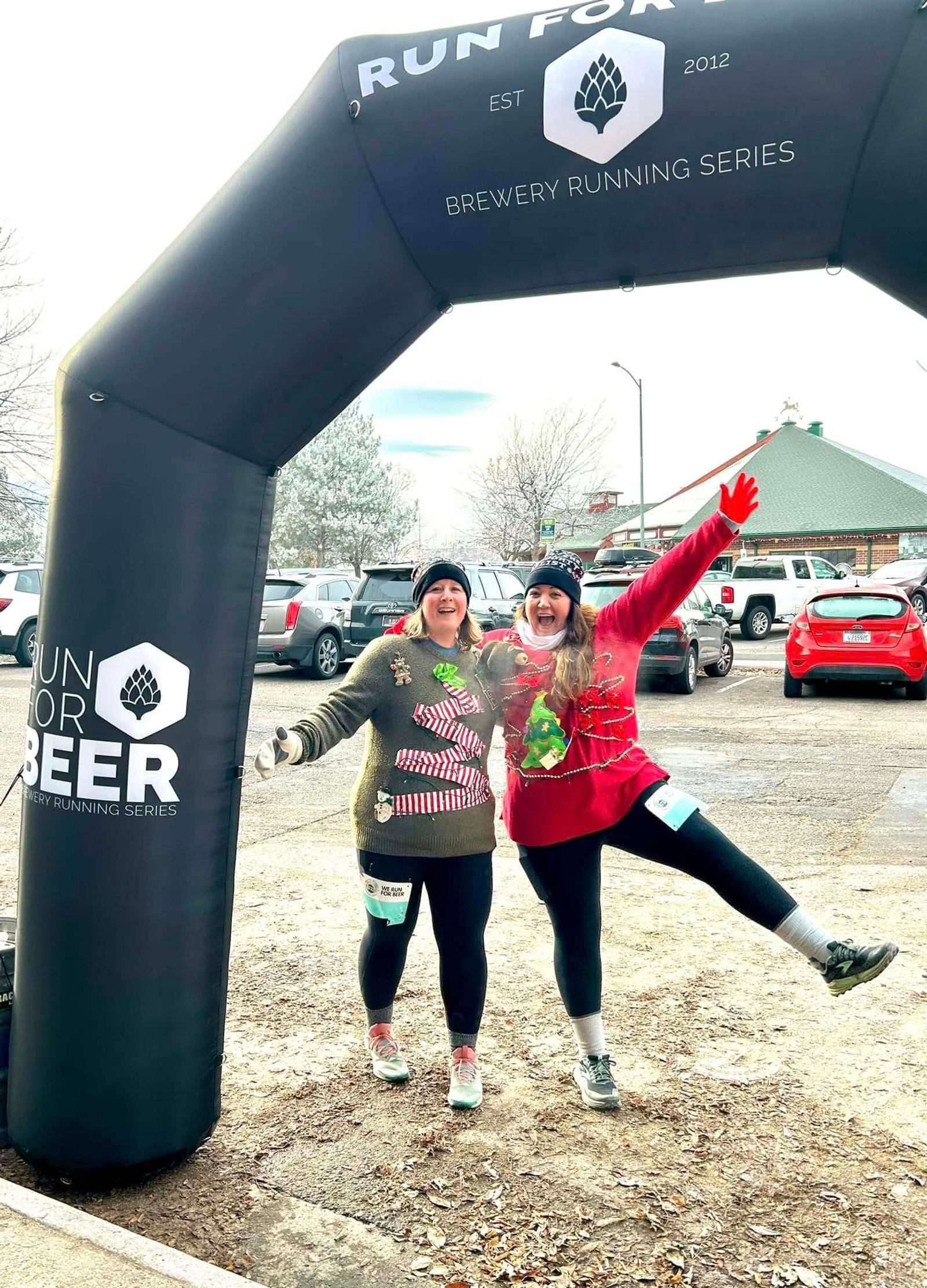 Two smiling people wearing festive sweaters and race bibs pose under an inflatable "Run for Beer" arch at an outdoor event, with snowy ground, parked cars, and trees in the background.