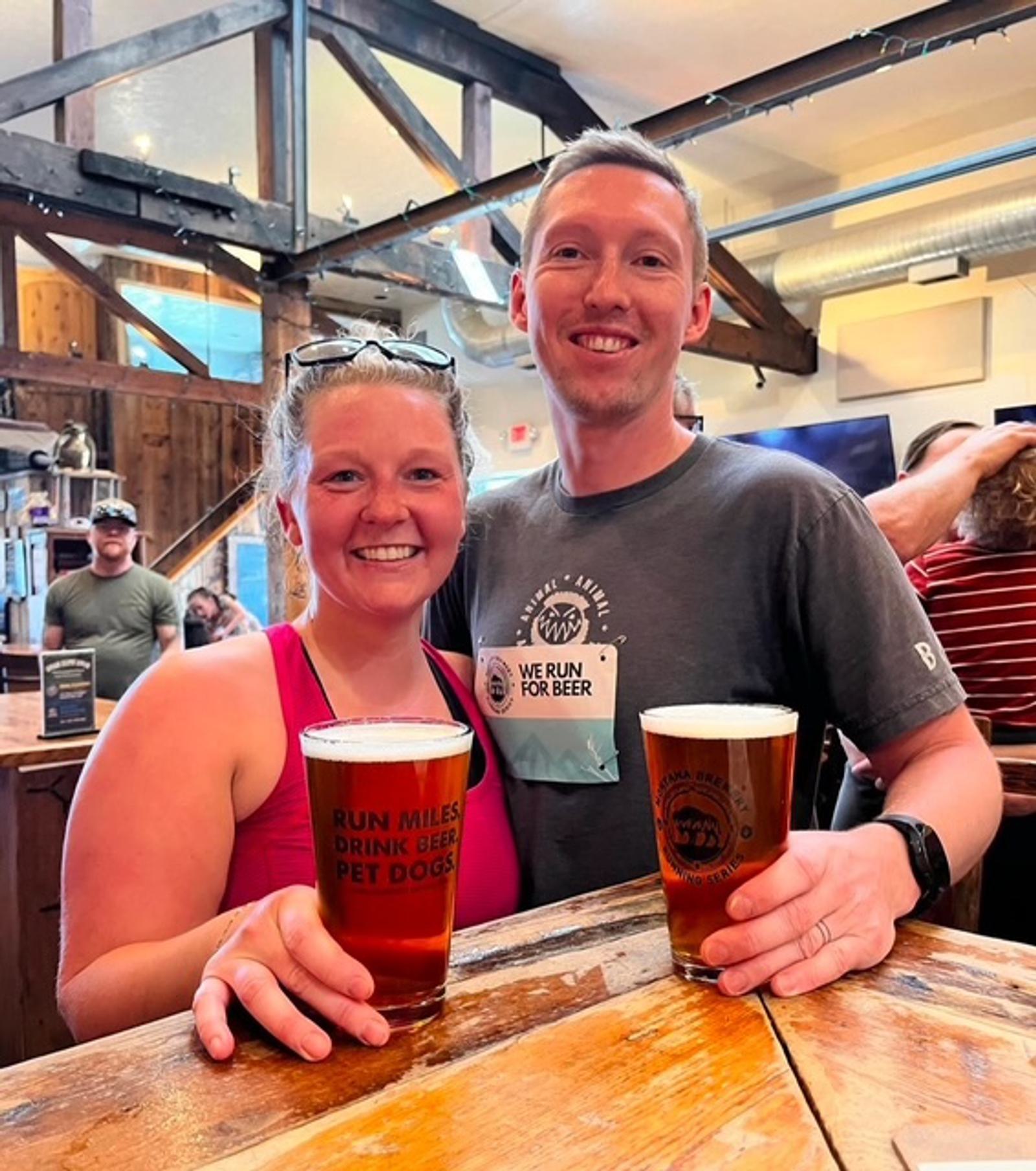 A smiling woman and man hold pints of beer at a wooden bar in a lively brewpub. The woman wears sunglasses on her head and a pink tank top; the man wears a gray shirt with a beer-themed logo. Other patrons are in the background.