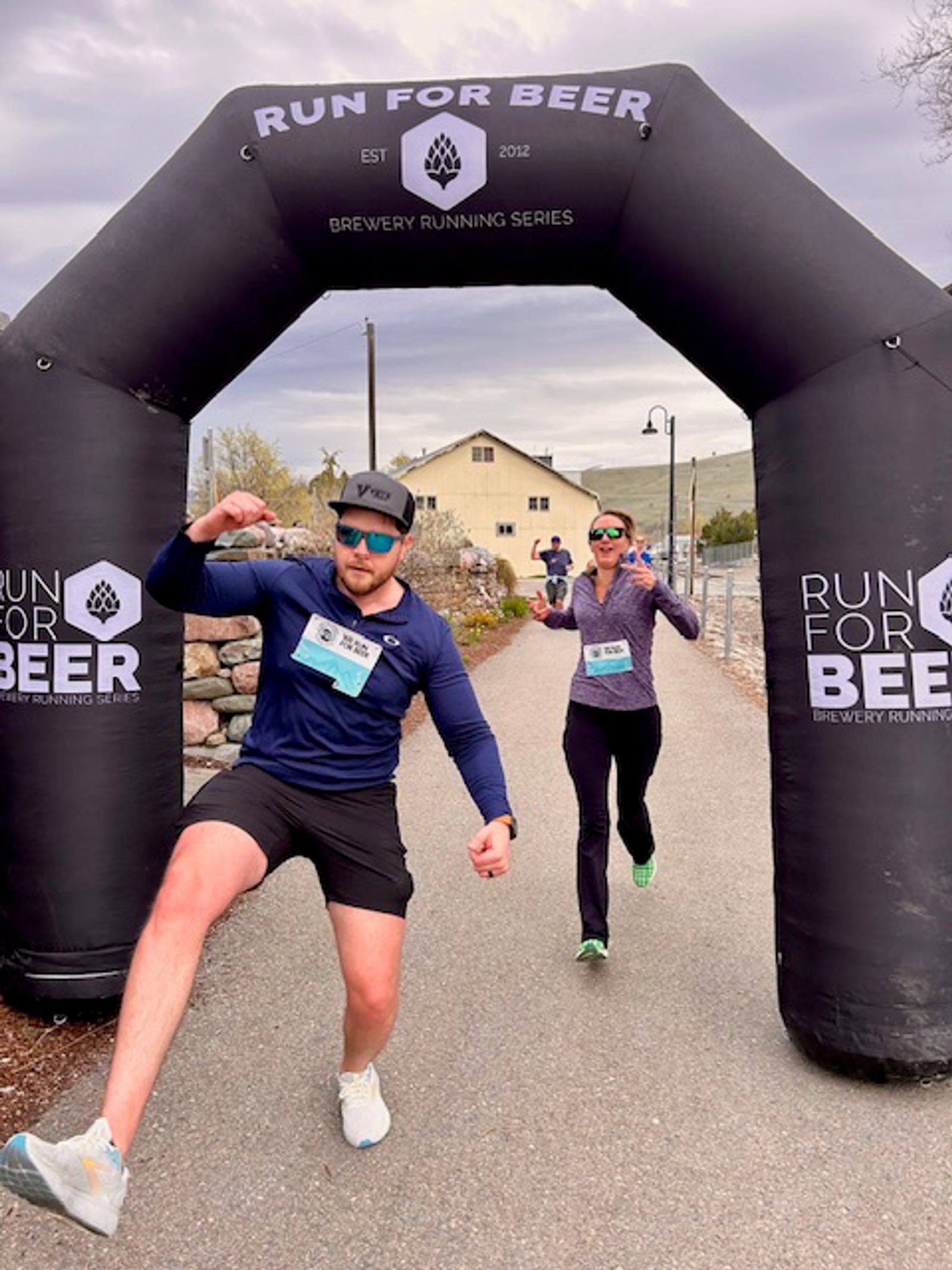 Two runners wearing race bibs and sunglasses cross the "Run For Beer" finish line arch. The man in front makes a playful pose, while the woman behind him smiles, both celebrating their race achievement.