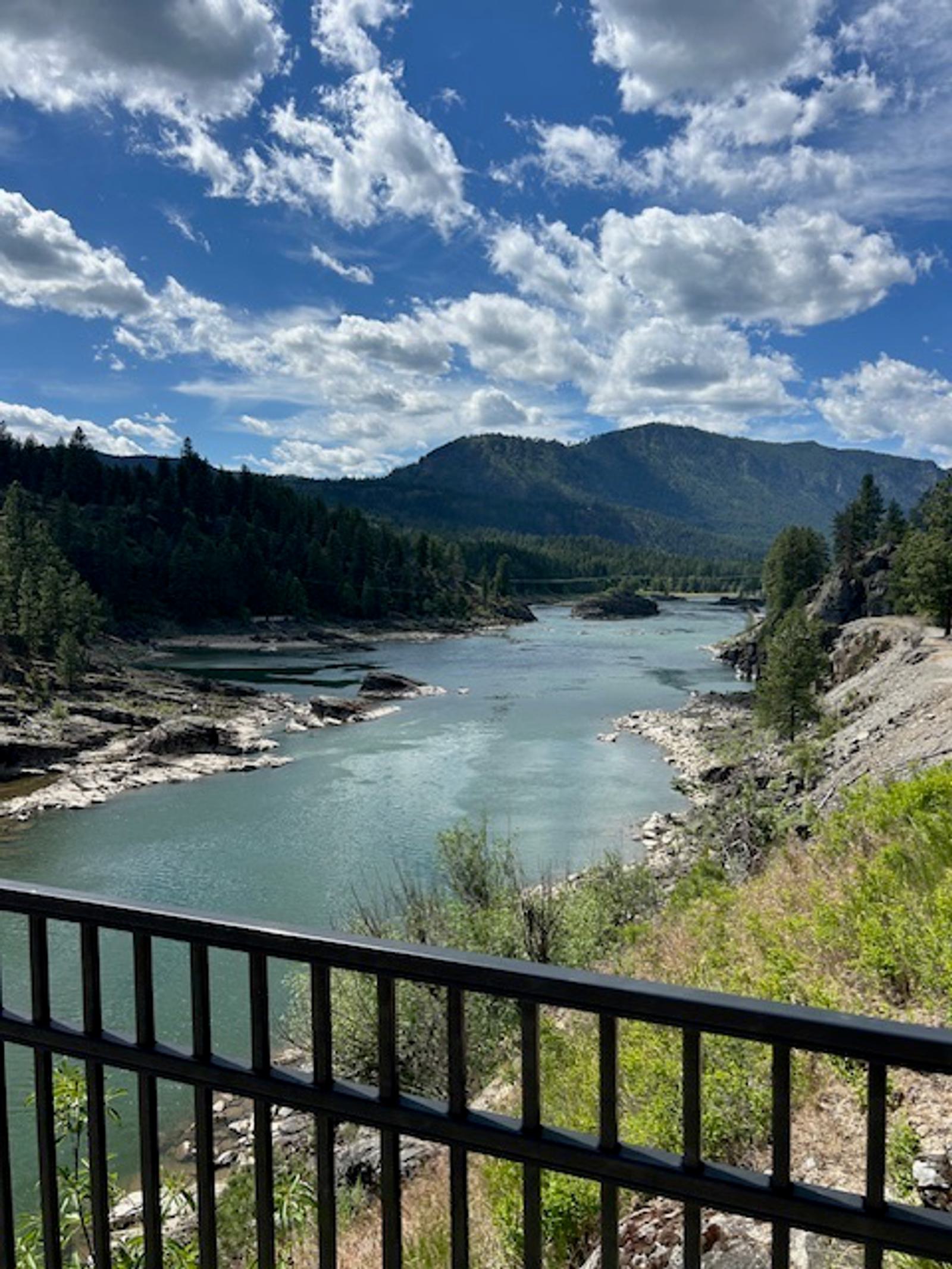 A scenic river flows between forested hills and mountains under a partly cloudy blue sky, viewed from behind a black metal fence in the foreground.