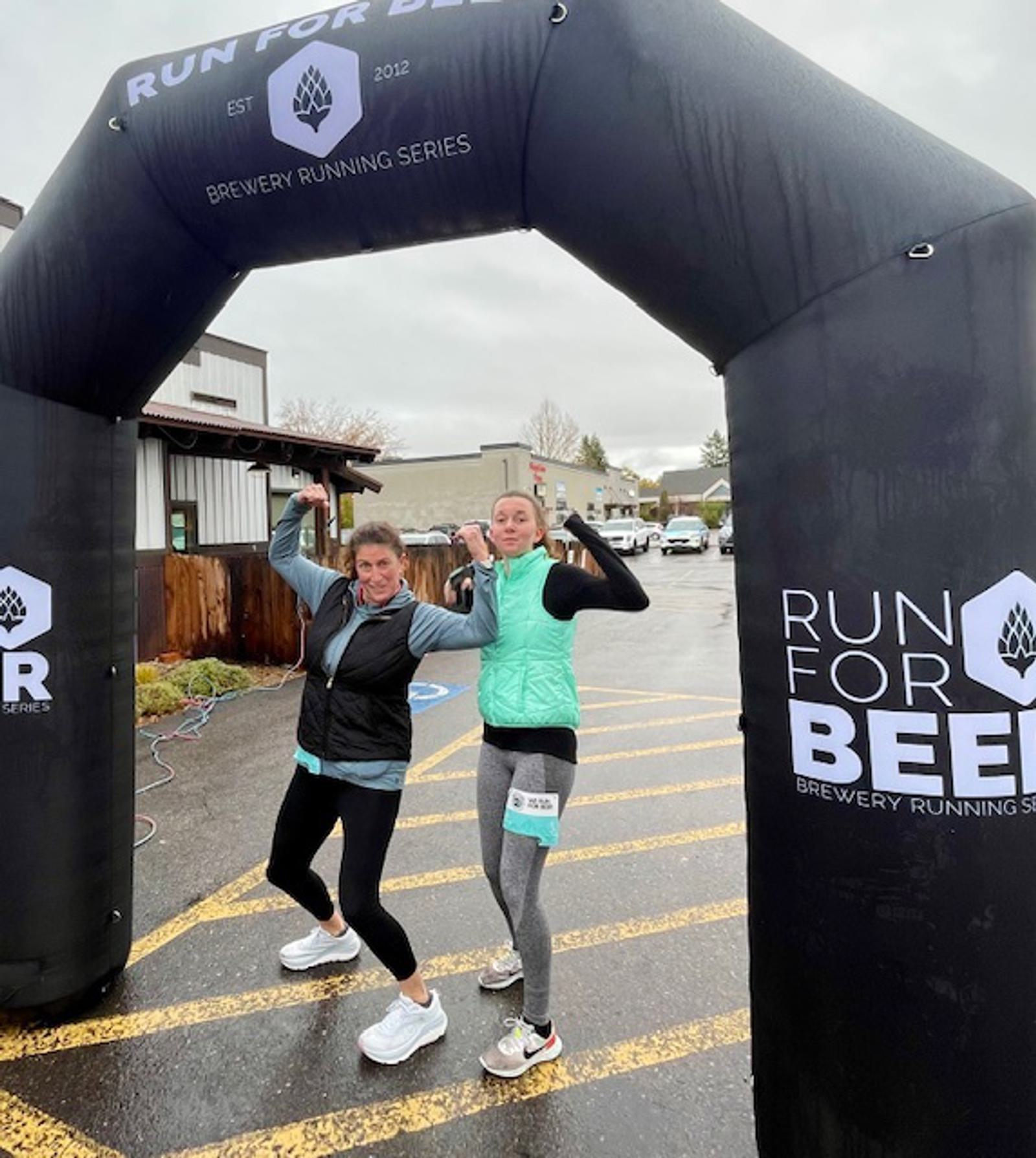 Two women in athletic wear pose and flex under an inflatable arch that reads "Run For Beer" at a Brewery Running Series event on a wet, overcast day. They appear happy and triumphant.