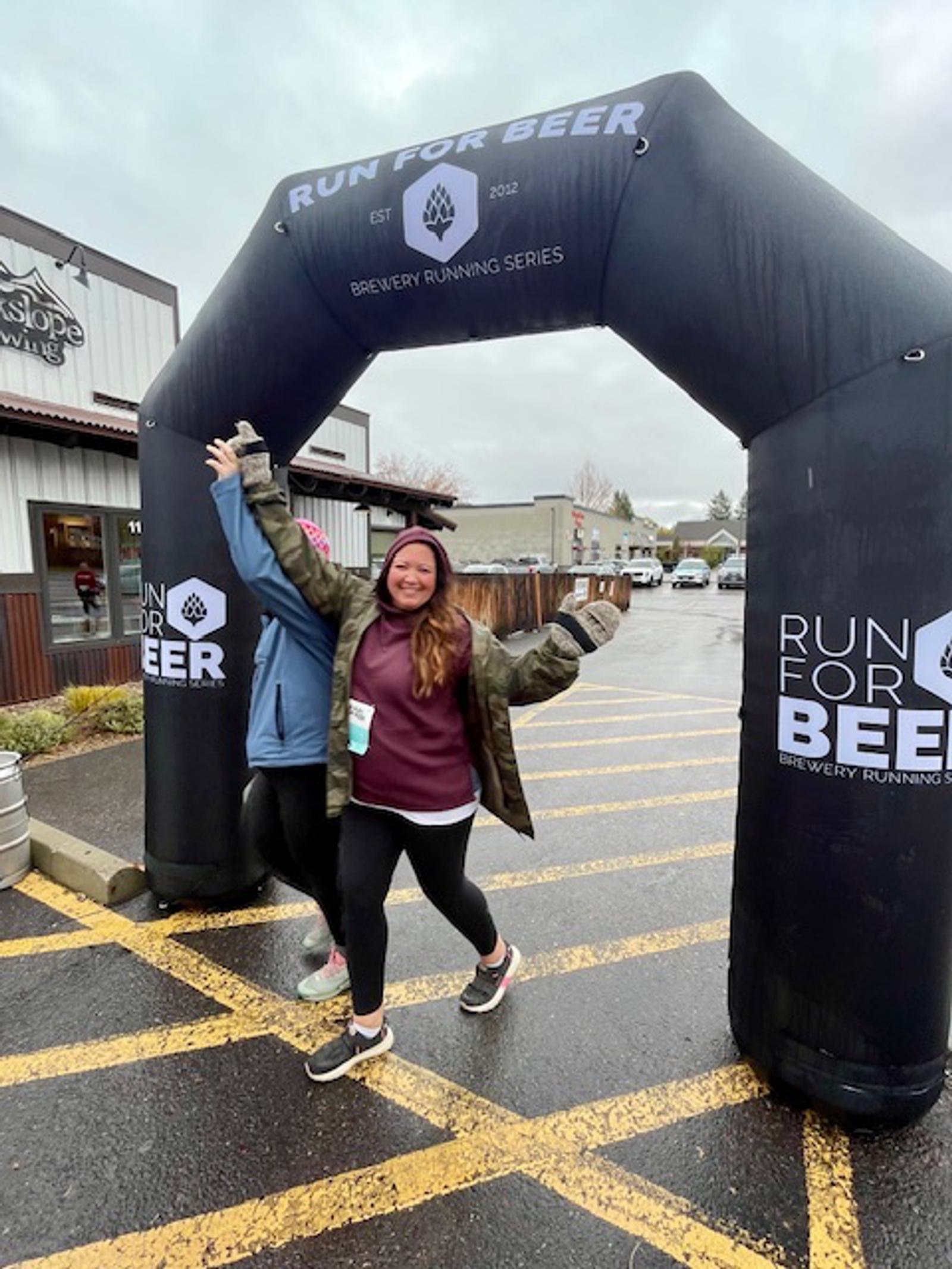 A smiling woman poses with arms outstretched under a black "Run for Beer" inflatable arch at a brewery running event. Another person stands behind her, partially hidden. It's a cloudy, rainy day.