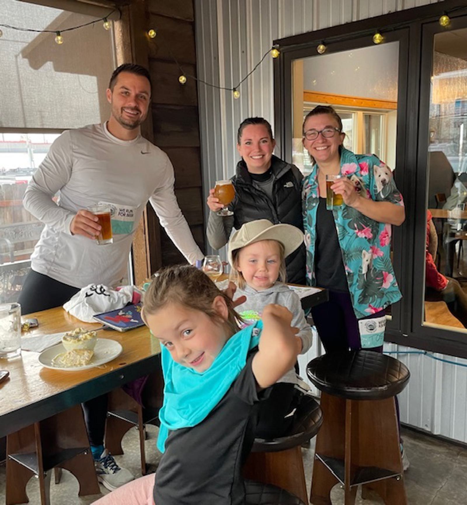 Three adults stand smiling with drinks behind two young children seated at a table with food. The group appears happy inside a cozy restaurant with string lights and large windows.