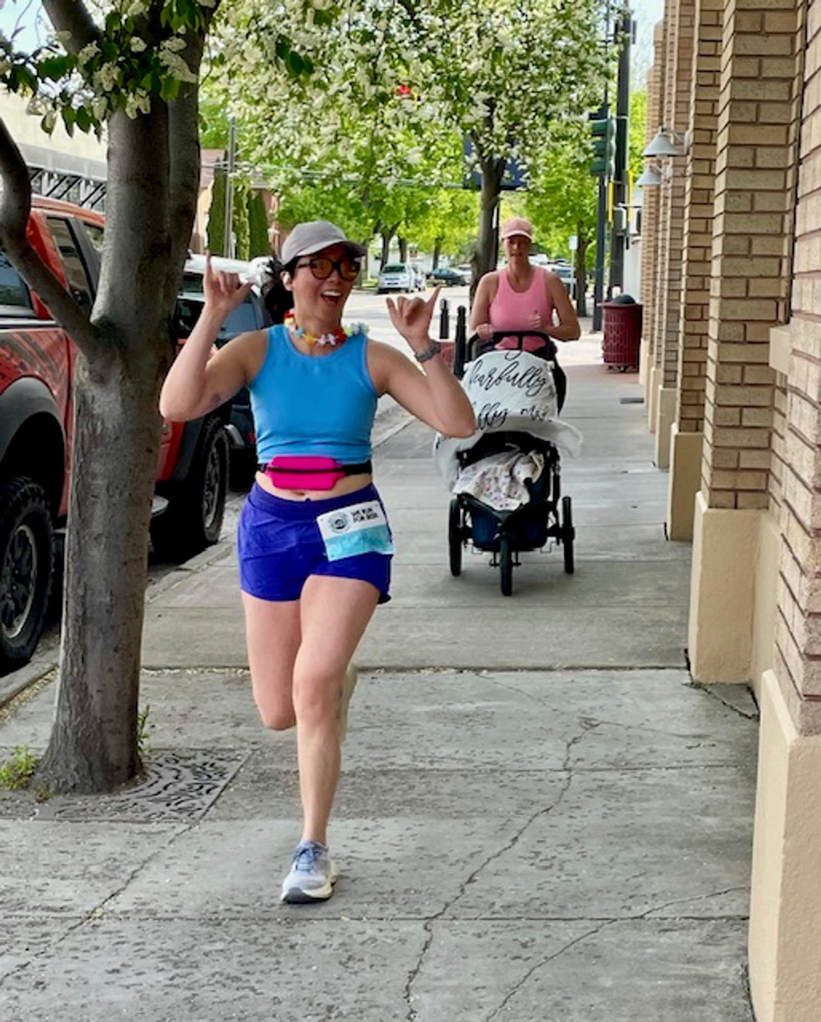 A woman in running gear smiles and gestures with both hands while jogging on a sidewalk. Behind her, another woman pushes a stroller. Trees and brick buildings line the street.