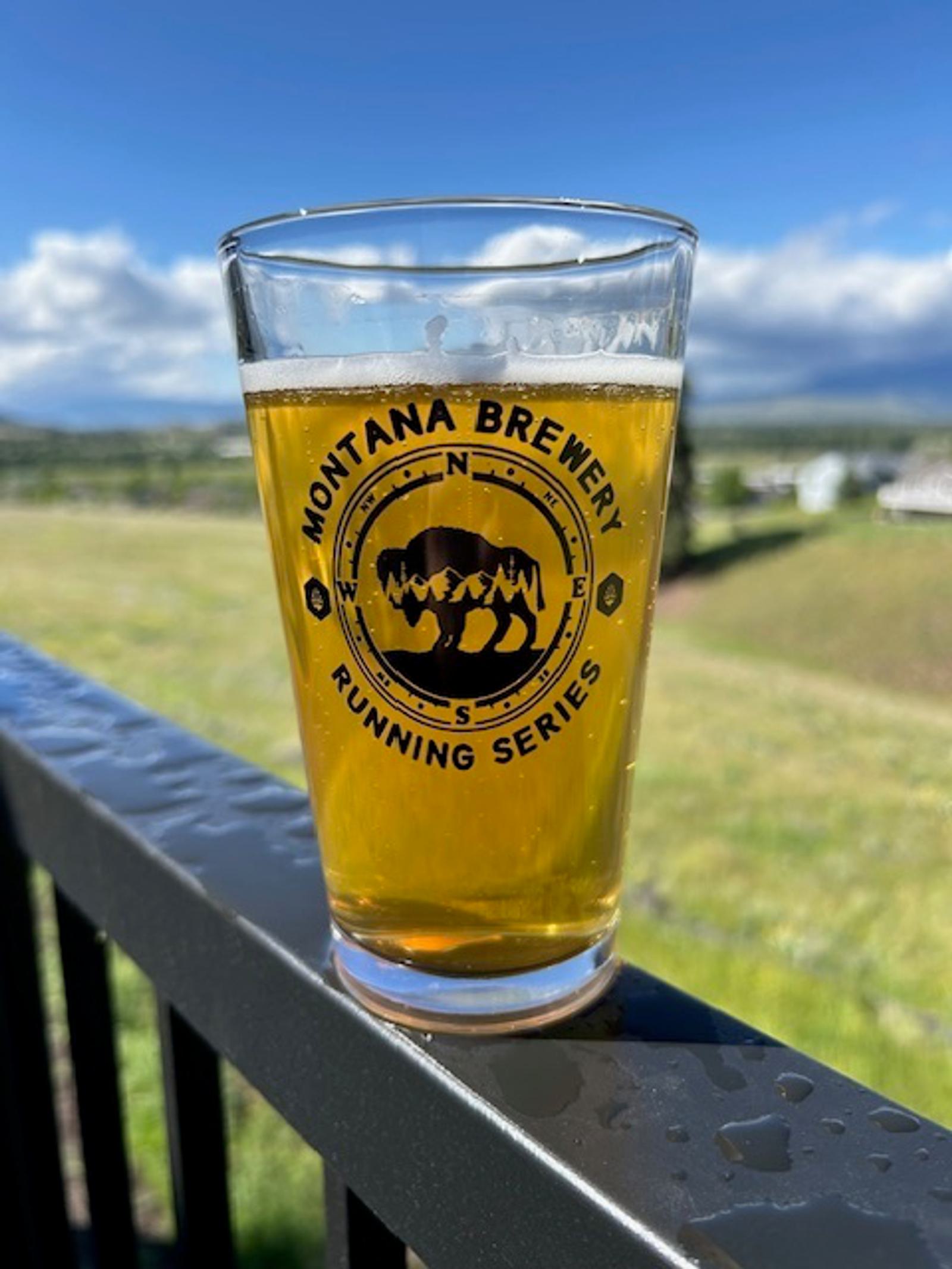 A pint glass of beer with "Montana Brewery Running Series" printed on it sits on a wet railing, overlooking a grassy landscape and a partly cloudy blue sky in the background.