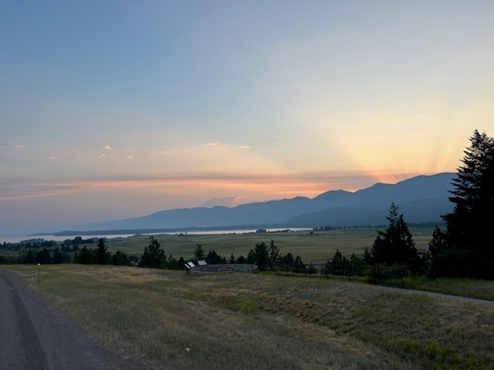 A scenic view of a rural road beside grassy fields at sunset, with mountains and a lake in the distance. Sun rays shine through the clouds, casting a soft glow over the landscape. Trees frame the scene on the right.