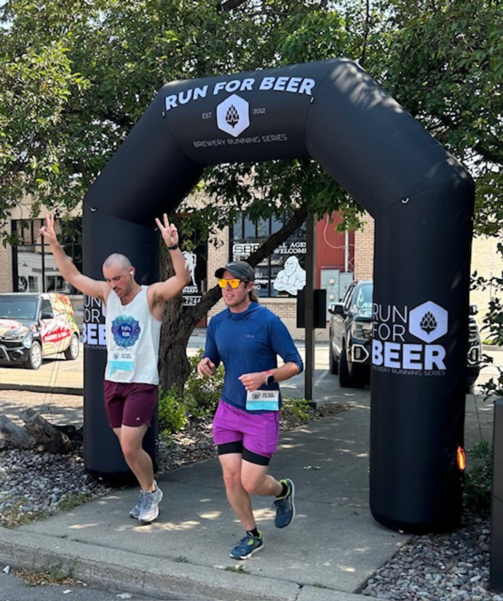 Two runners cross a "Run For Beer" finish arch. One smiles and raises both arms in a peace sign, while the other runs beside him, both wearing race bibs and athletic gear. Trees and parked cars are visible in the background.