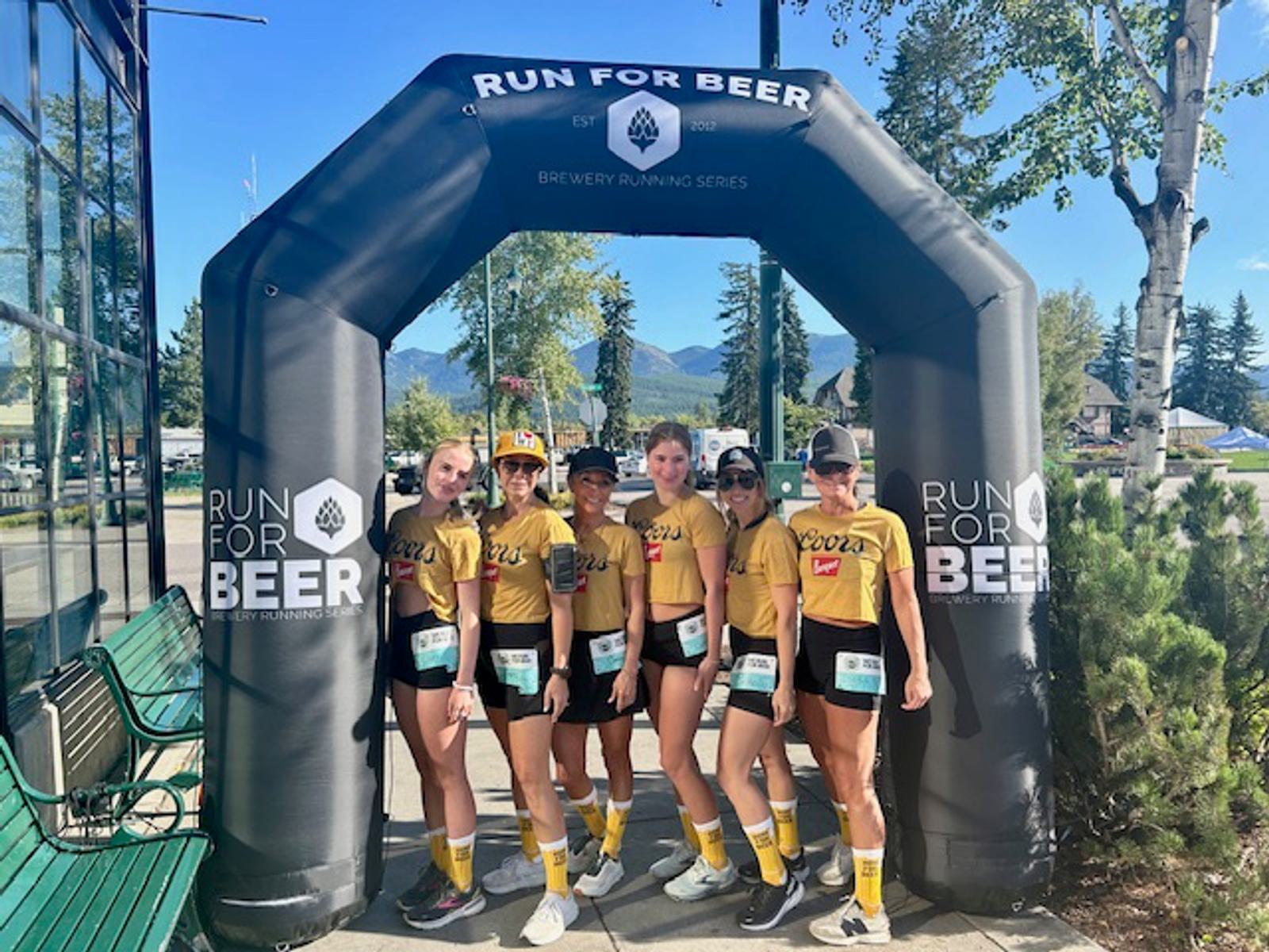 Six women in matching yellow shirts and black shorts stand under a “Run for Beer” inflatable arch outdoors, posing for a photo on a sunny day with mountains and trees in the background.