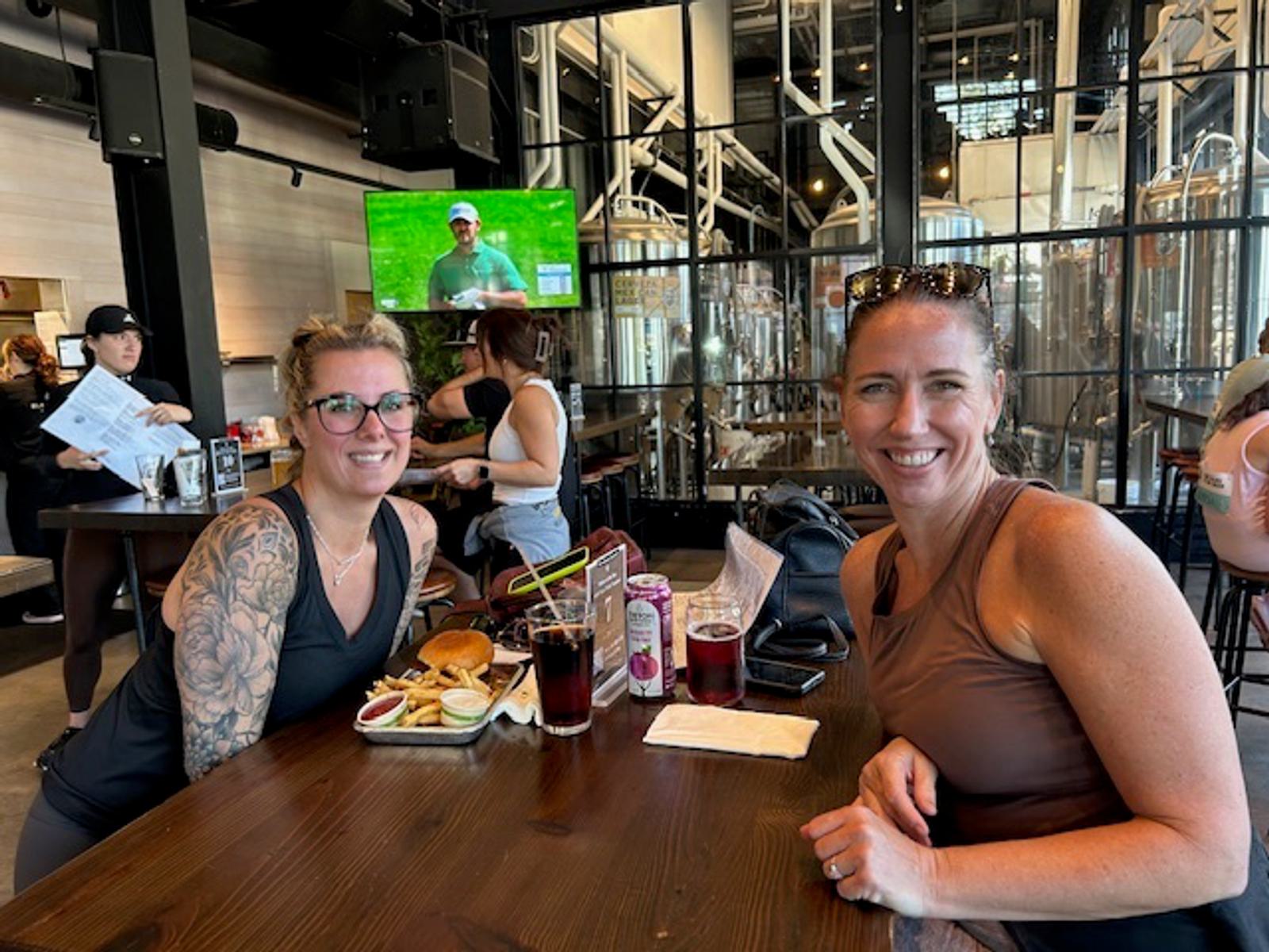 Two women sit smiling at a restaurant table with food and drinks. Behind them are large brewing tanks and other diners. A TV in the background displays a sports event. The setting is casual and bright.