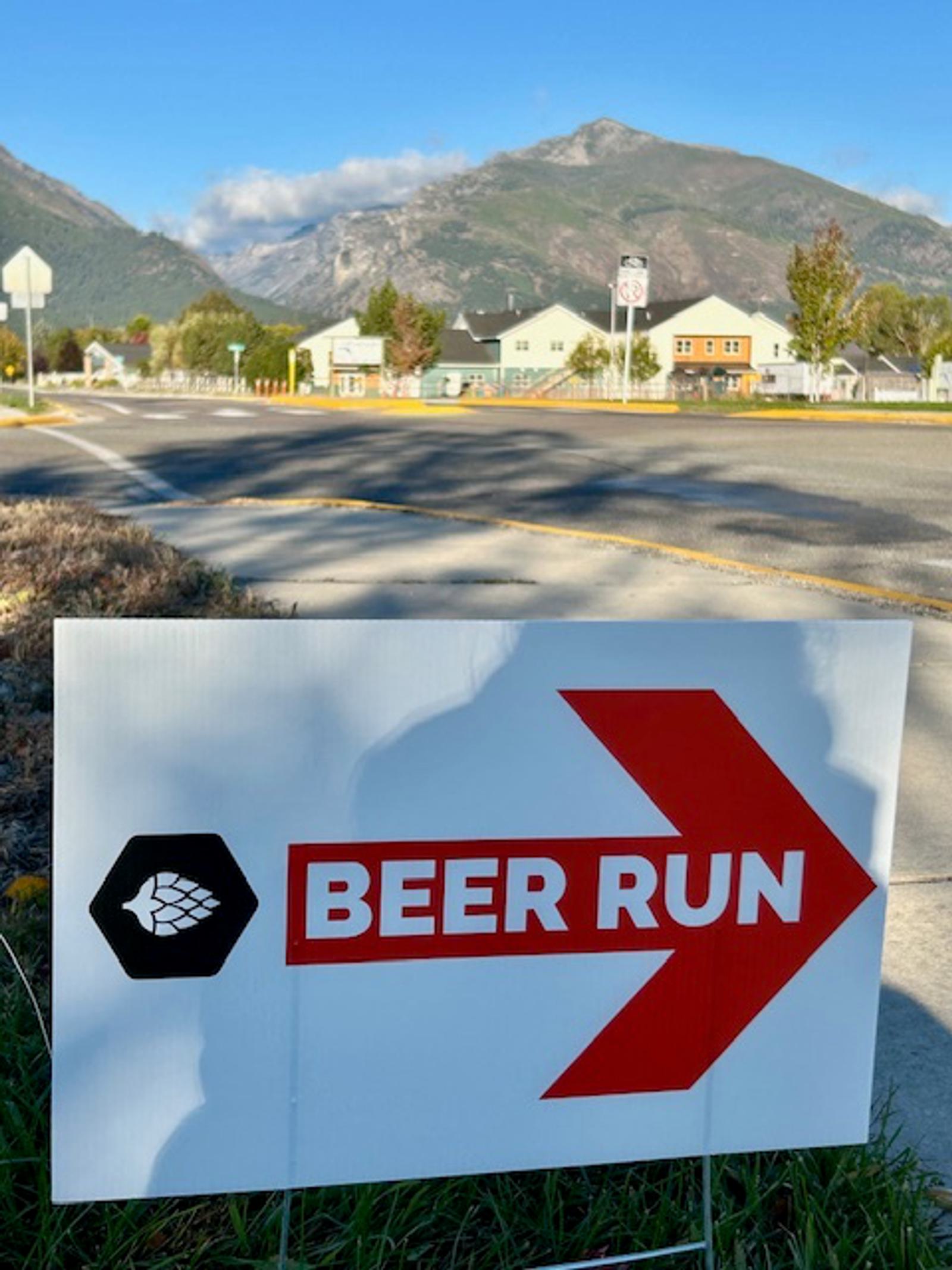 A yard sign with a red arrow and the words "BEER RUN" points right on a suburban street, with mountains, houses, and a blue sky in the background.