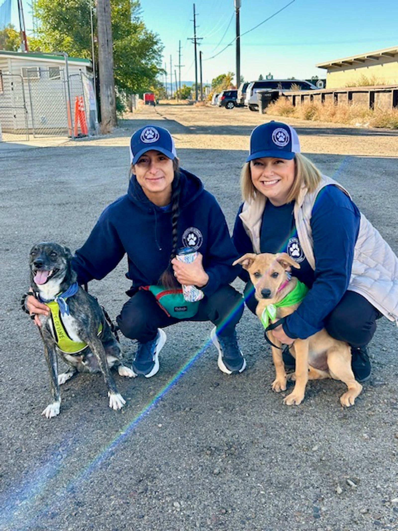 Two women in matching navy hats and sweatshirts kneel outdoors, each holding a dog on a leash. One dog is brindle with a yellow harness, the other is tan with a green bandana. Both women are smiling.