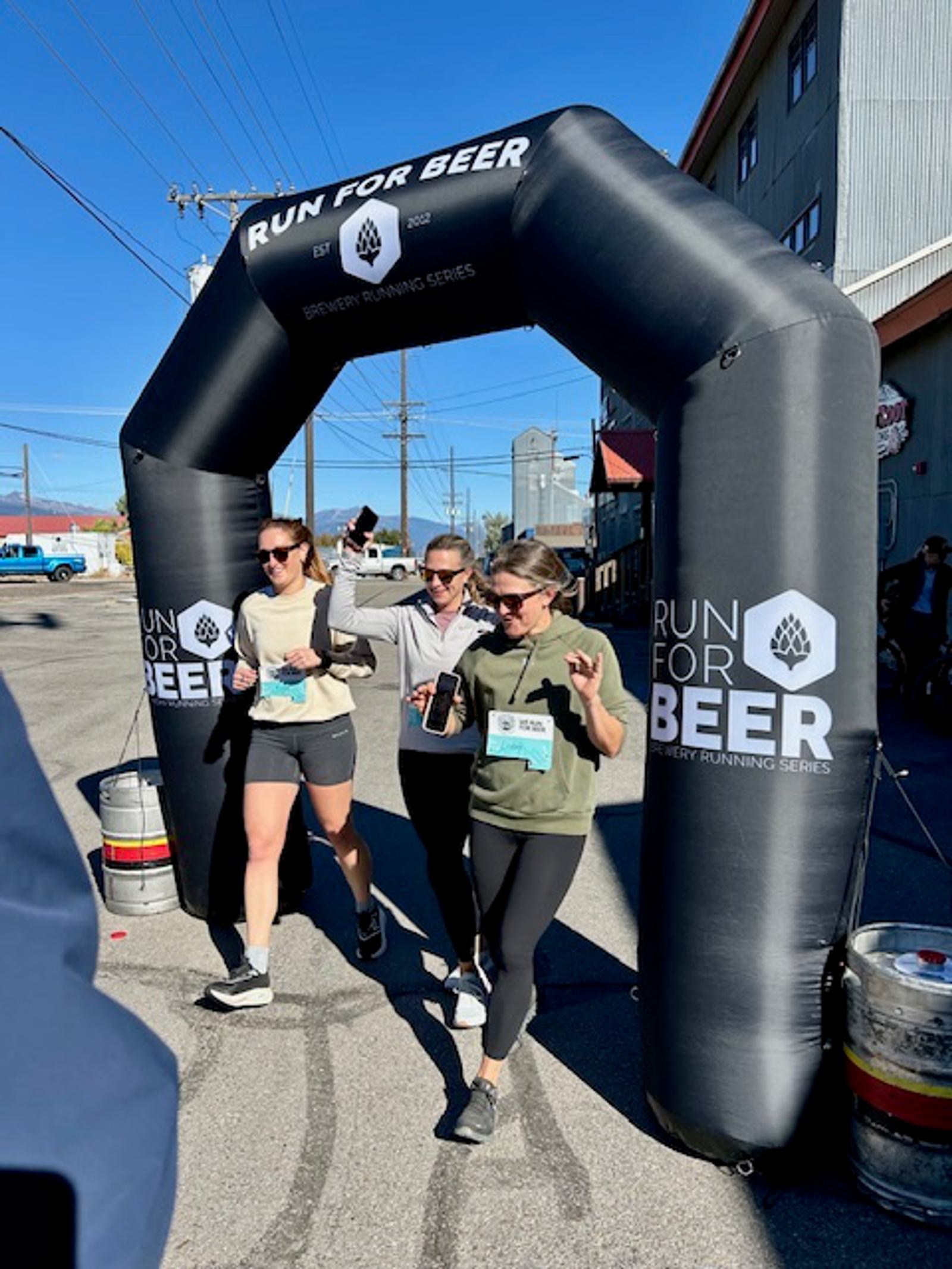 Three women wearing athletic clothing walk together under a black inflatable finish arch that reads "Run for Beer." Two hold drinks, and all appear cheerful. The event takes place outdoors on a sunny day next to buildings.