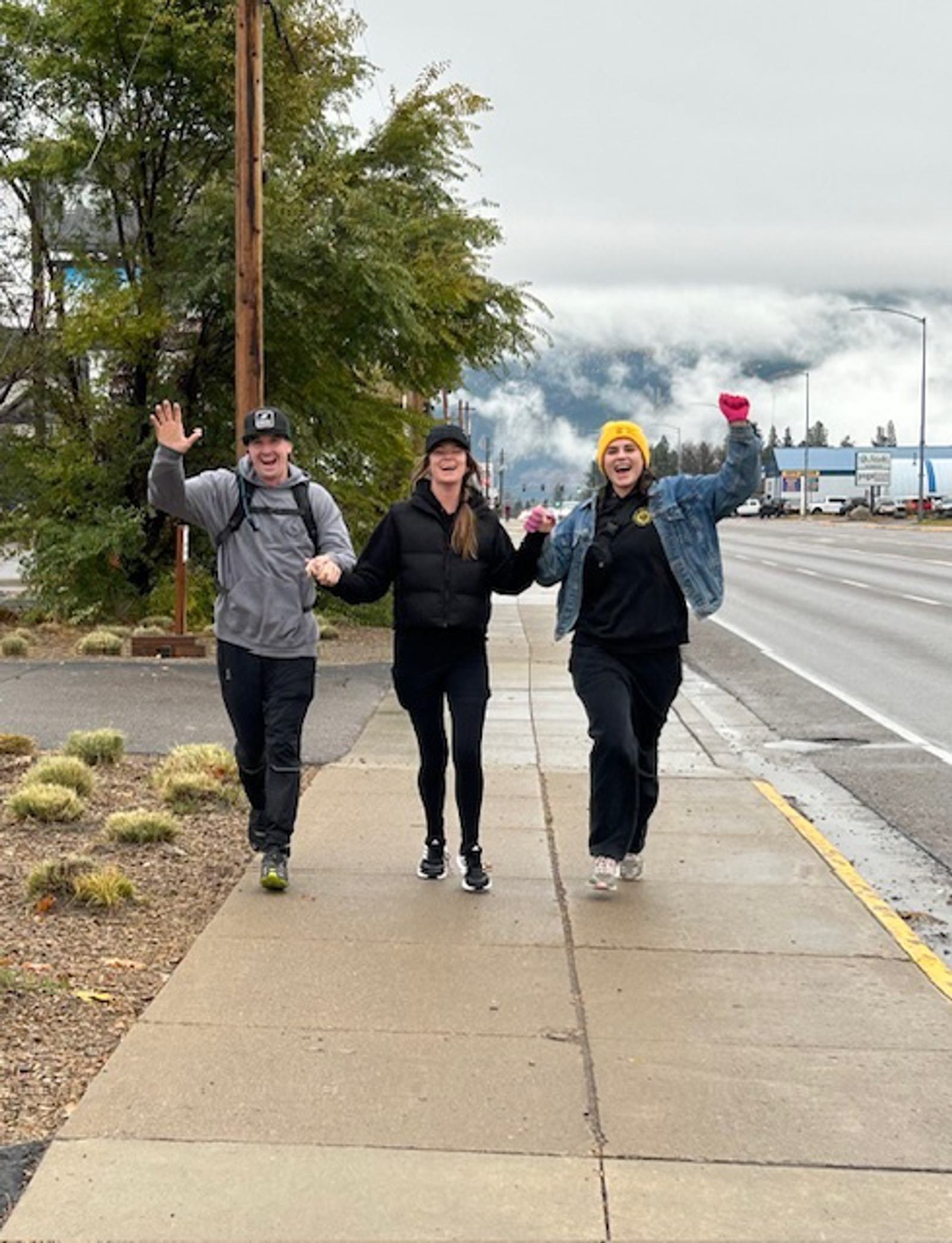 Three people walk and smile on a wet sidewalk by a road, wearing casual clothes and hats or a beanie. They hold hands and wave, with cloudy skies and mountains visible in the background.