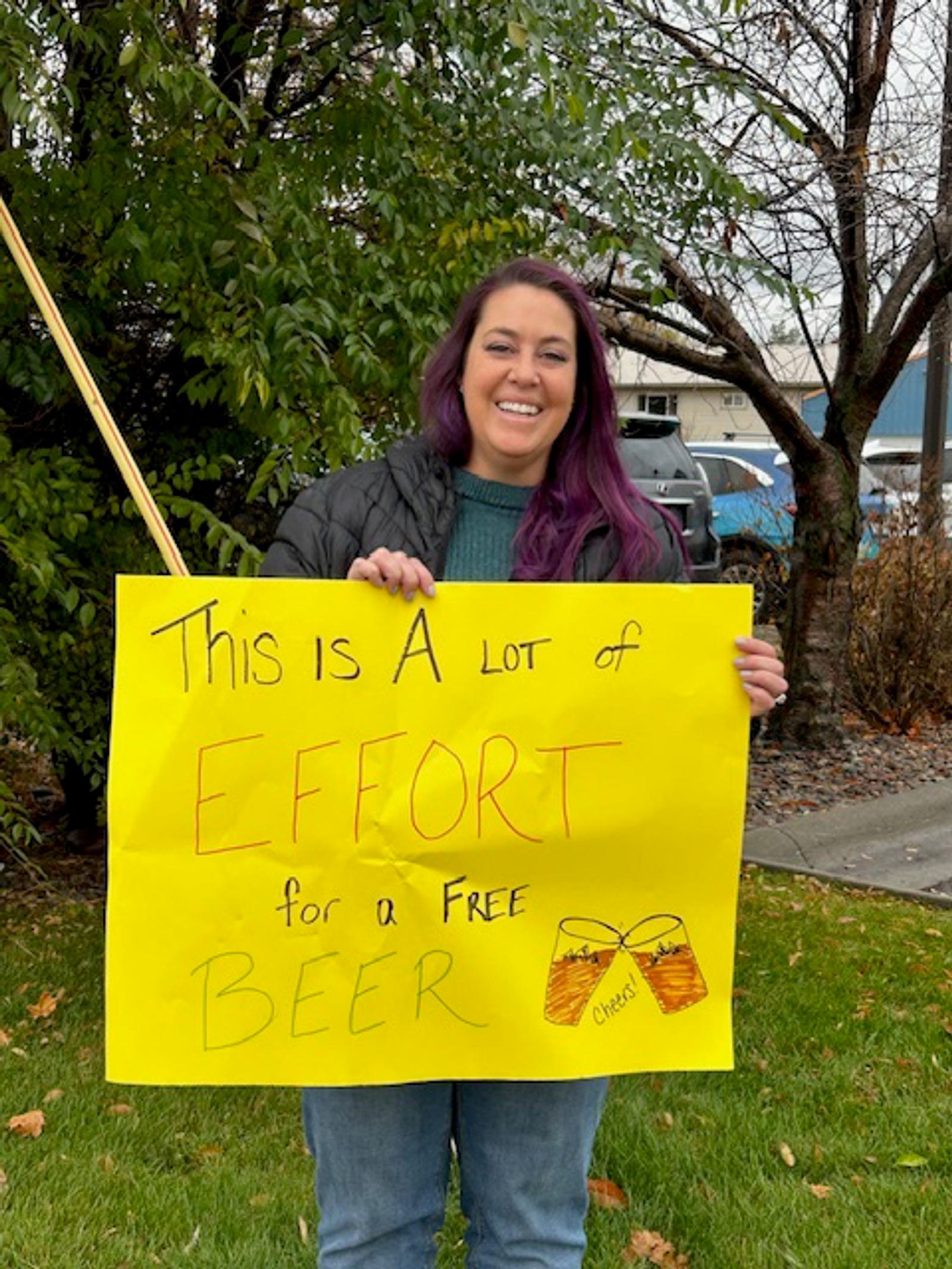 A smiling woman with long purple hair holds a bright yellow sign that says, "This is a lot of effort for a free beer," with two drawn beer mugs at the bottom. She stands outside on grass near trees.