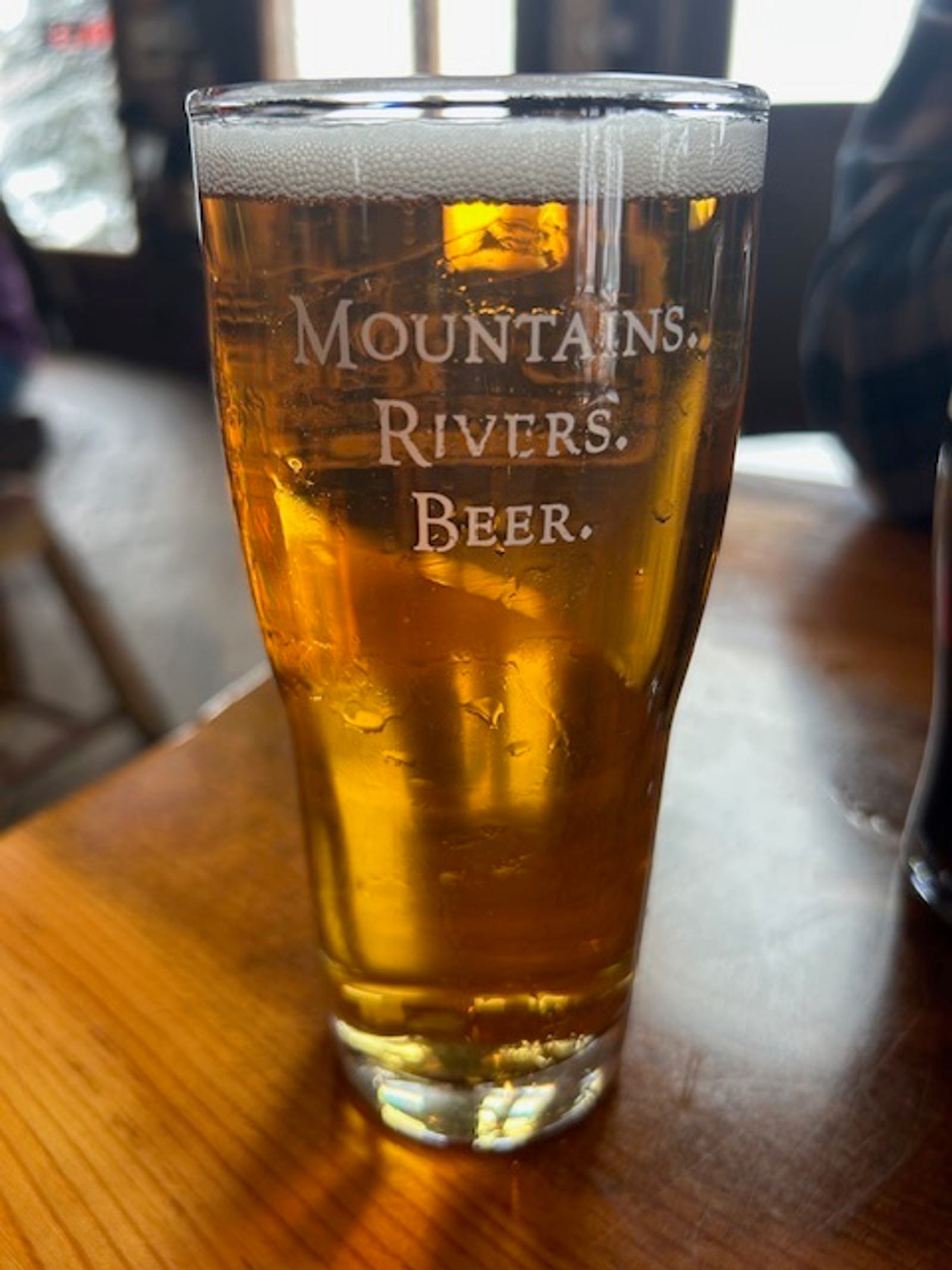 A pint glass of golden beer on a wooden table, with the words "MOUNTAINS. RIVERS. BEER." etched on the glass. Light shines through the beer, and the background is softly blurred.