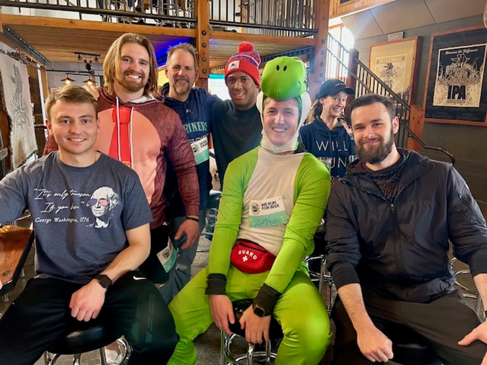 A group of six smiling adults sit together indoors. One person is dressed in a green dinosaur costume with a tail and hood. The others wear casual shirts and jackets. The background shows wooden beams and framed artwork.