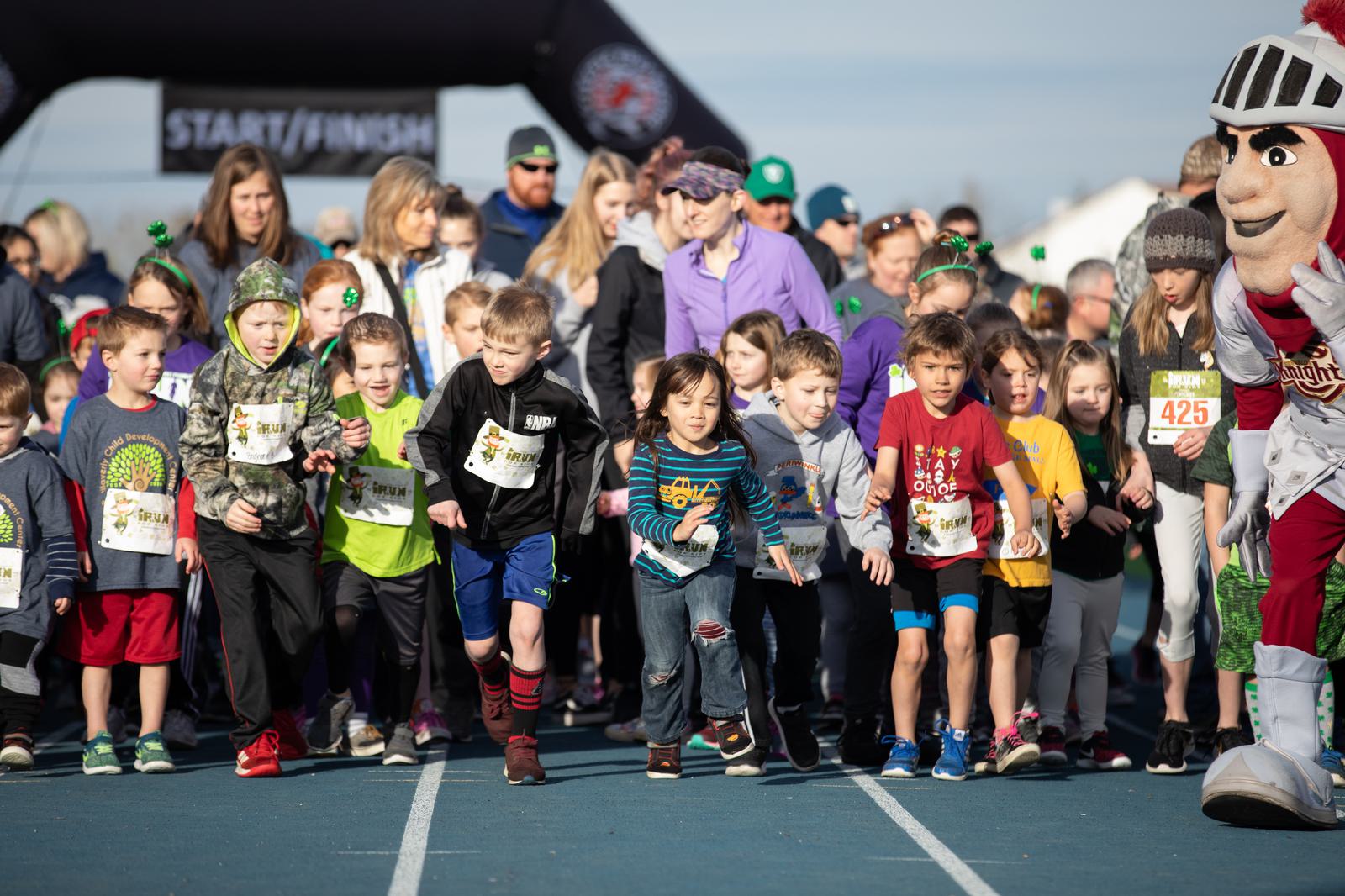 A group of children wearing athletic gear start running at a race event. Adults and a mascot stand behind them on a track, with a start/finish line arch in the background. The sky is clear, and it appears to be a sunny day.