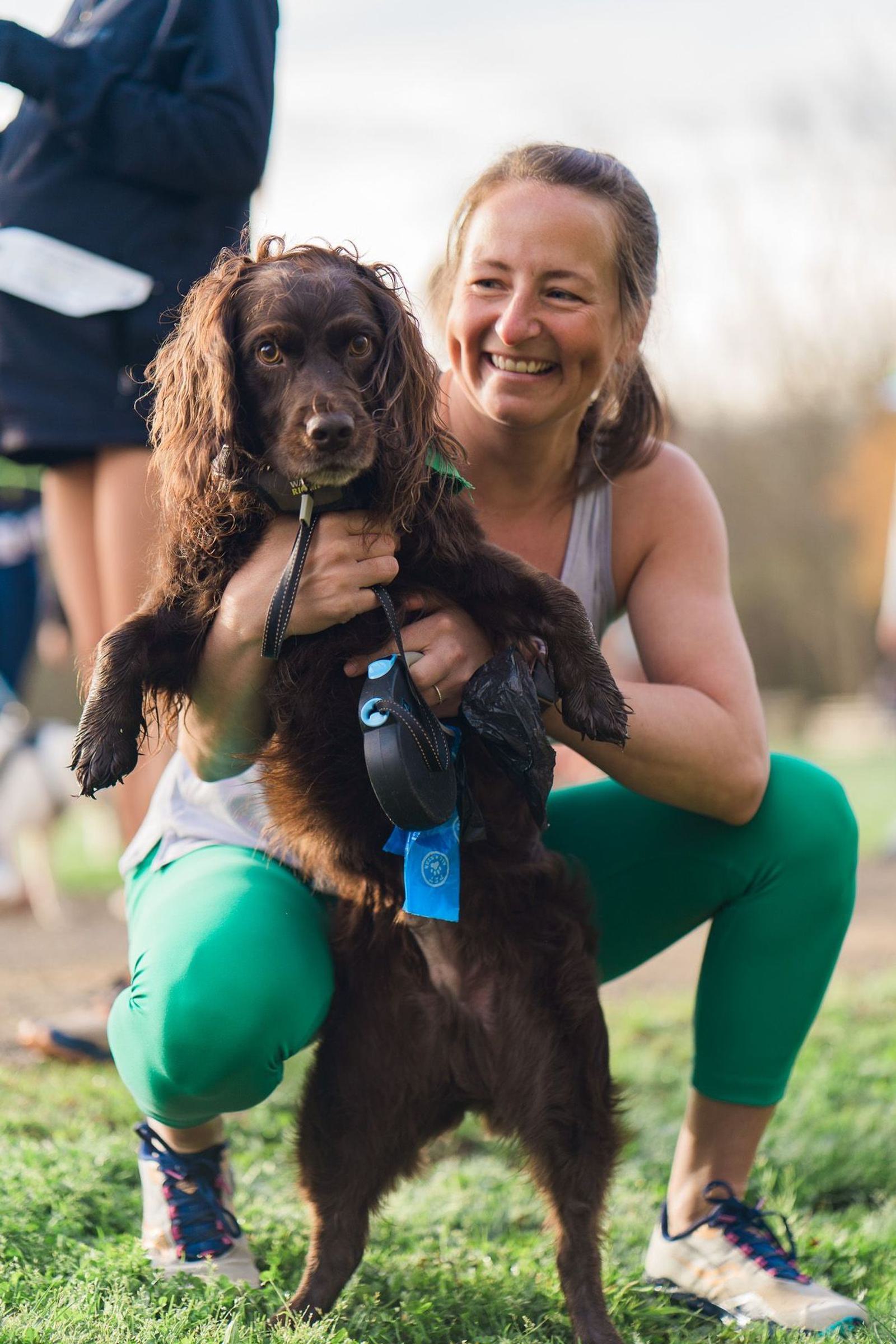 A smiling woman crouches on the grass, holding a brown, curly-haired dog with a blue ribbon. The dog wears a collar and leash. Other people are visible in the blurry background, suggesting an outdoor event.