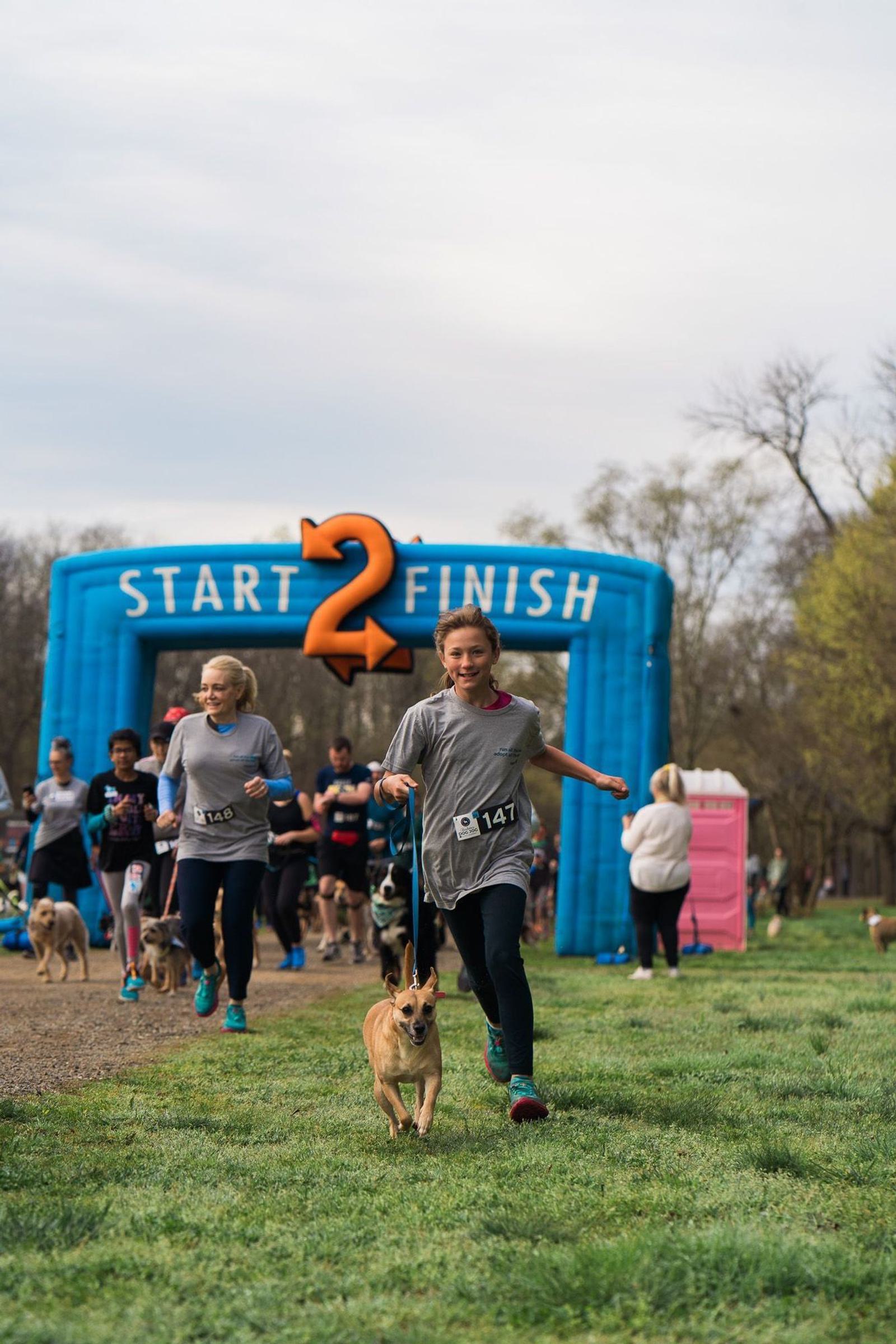 A smiling young girl runs energetically with her dog near the finish line of a race event. The blue inflatable archway reads "Start 2 Finish." Participants and spectators are visible in the background on a grassy path.