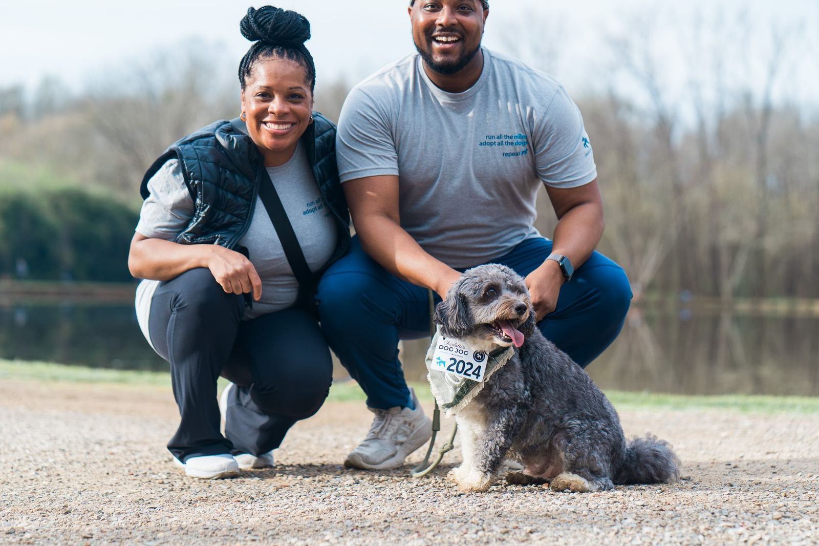 Two people are smiling and crouching beside a small dog wearing a race bib numbered 2024. They are outdoors on a gravel path near a body of water, with trees in the background.