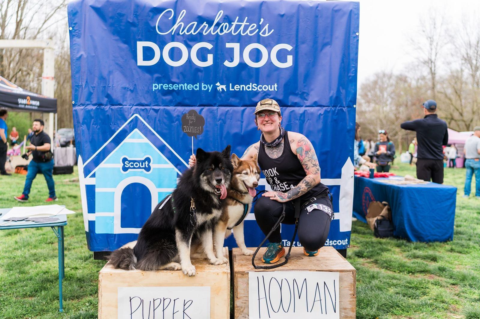 A person kneels beside two dogs on podium boxes labeled "Pupper" and "Hooman" at an outdoor event. A large banner behind them reads "Charlotte's DOG JOG presented by LendScout." Other participants and booths are visible in the background.