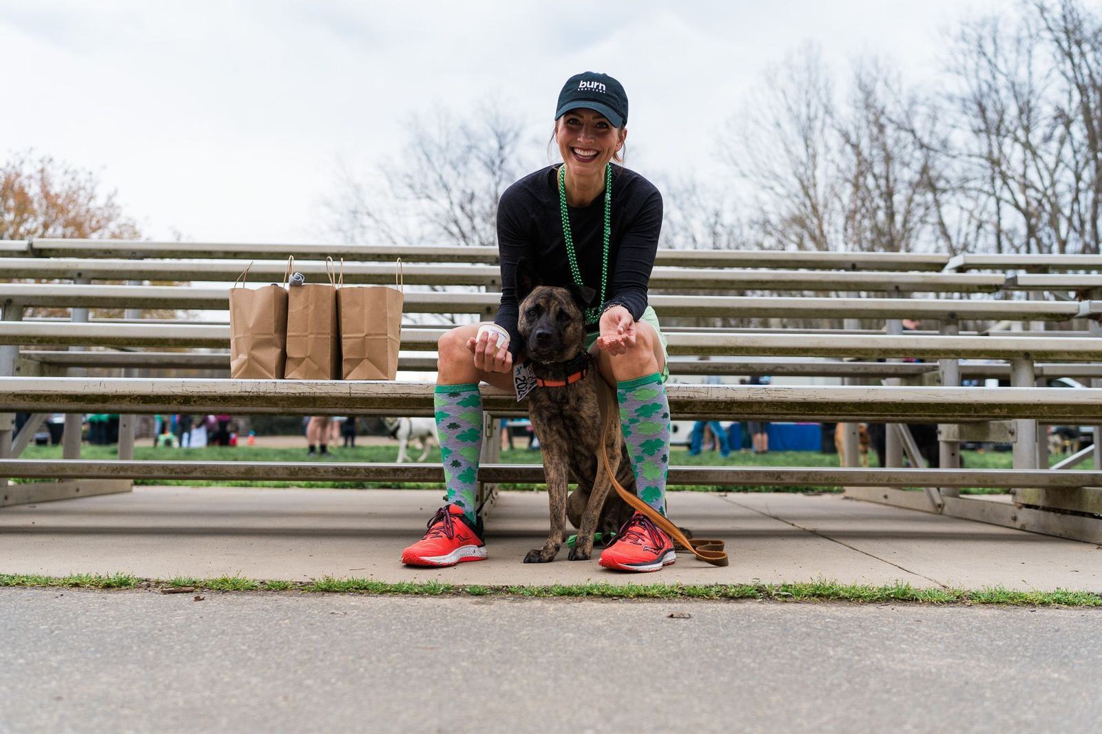 A person wearing a black cap, a black shirt, colorful knee socks, and red sneakers is sitting on bleachers with a brown dog. They are both looking at the camera. There are brown paper bags on the bench beside them, and trees are visible in the background.