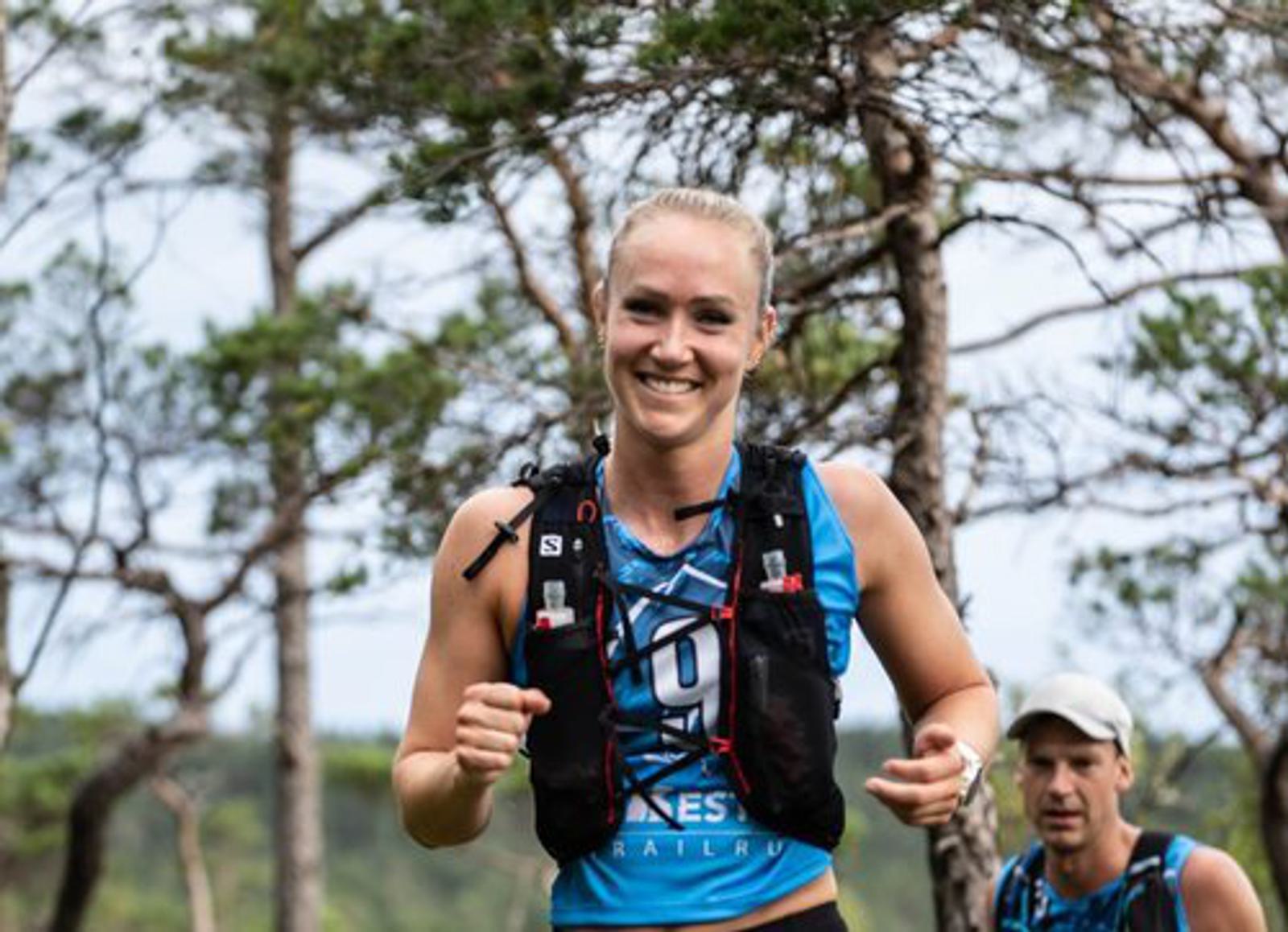 A person running outdoors on a trail, wearing a blue top and a hydration vest. They appear to be smiling, surrounded by trees. Another person is visible in the background, wearing a cap.