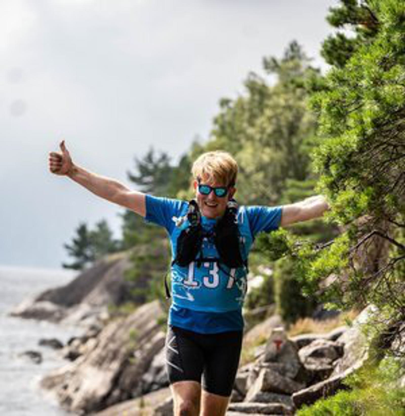 A person in a blue sports jersey and sunglasses jogs along a rocky coastal path lined with trees, giving a thumbs-up. The background features a cloudy sky and a view of the water.