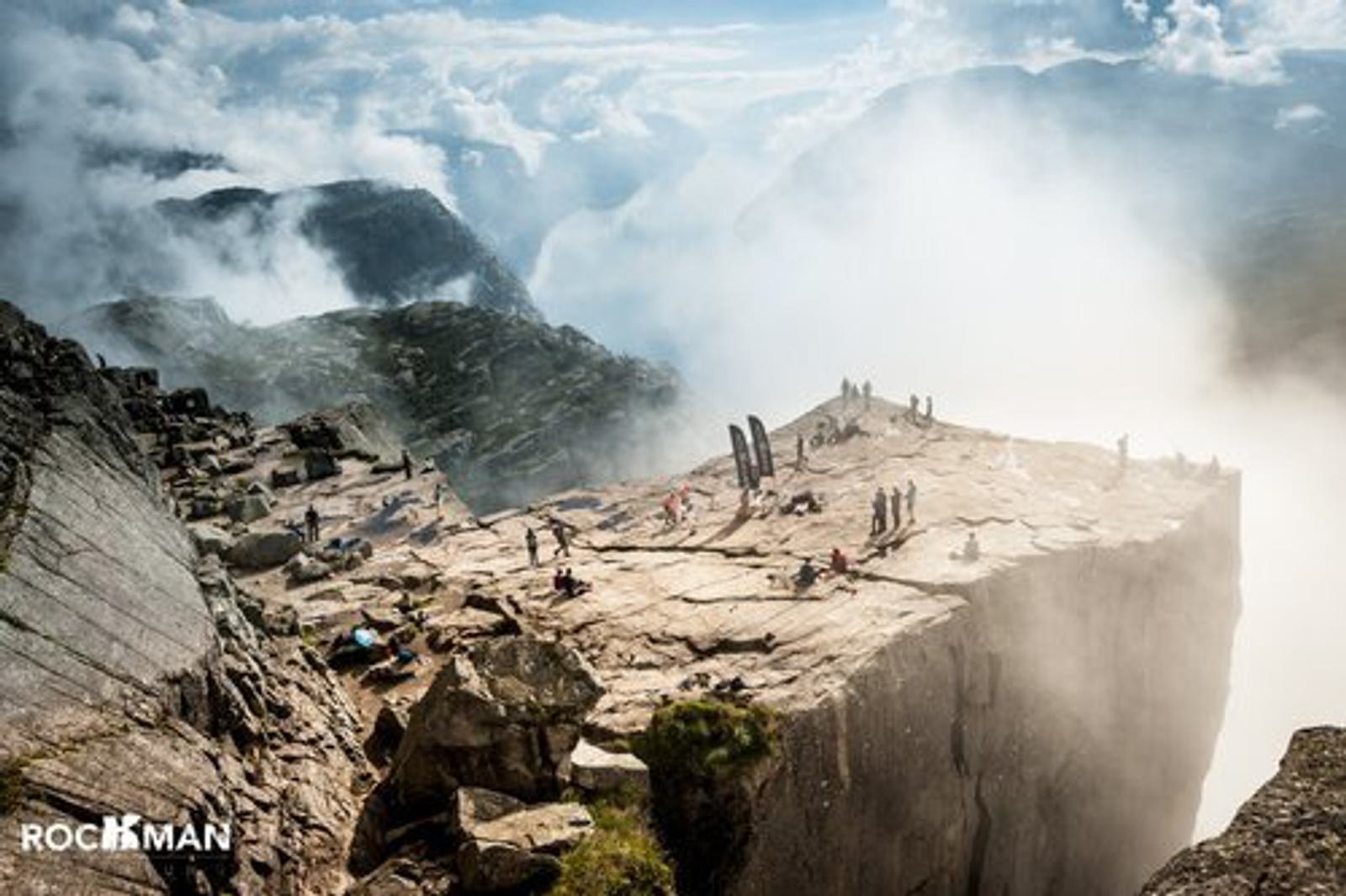 A breathtaking view of people standing and walking on the edge of a large, flat rock formation surrounded by misty clouds and mountainous landscape. The sky is partly cloudy, creating a dramatic atmosphere.