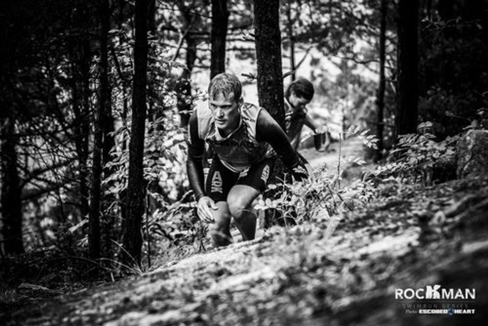 Two athletes run uphill through a forest trail. The leading runner looks focused as he ascends a rocky path surrounded by trees. The black and white image emphasizes the intensity and challenge of the race.