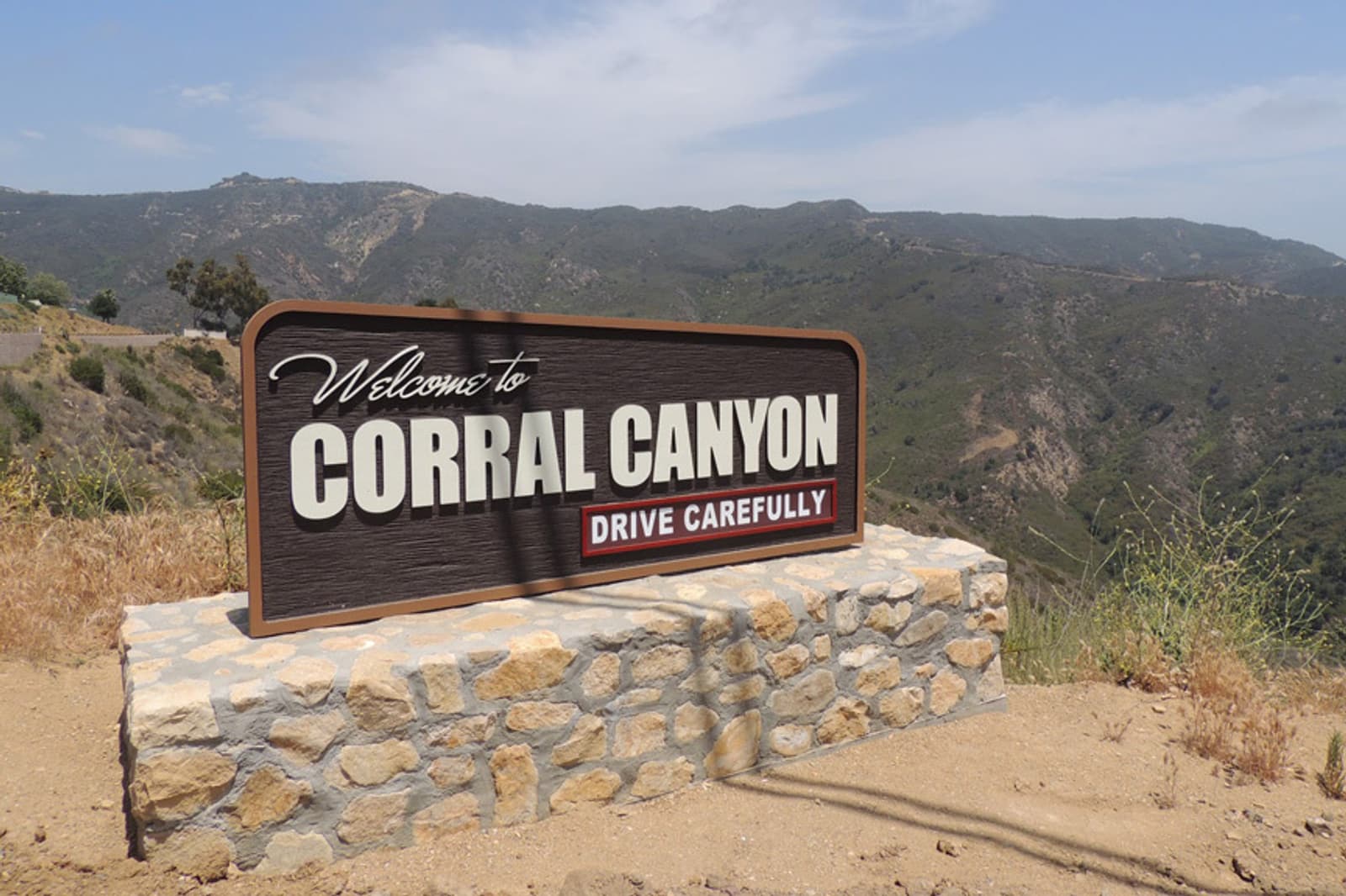 A sign that reads "Welcome to Corral Canyon Drive Carefully" stands on a stone base with a backdrop of rolling hills under a clear blue sky.