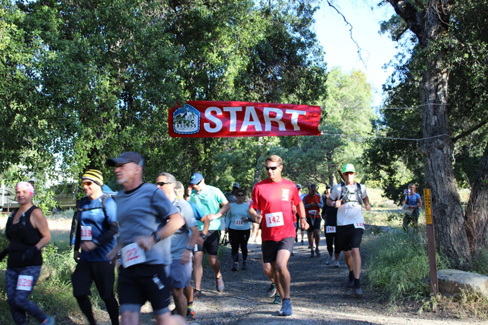 A group of runners in a race is starting under a red "START" banner. They are running on a dirt path surrounded by trees, with some participants wearing numbered bibs. It's a sunny day.