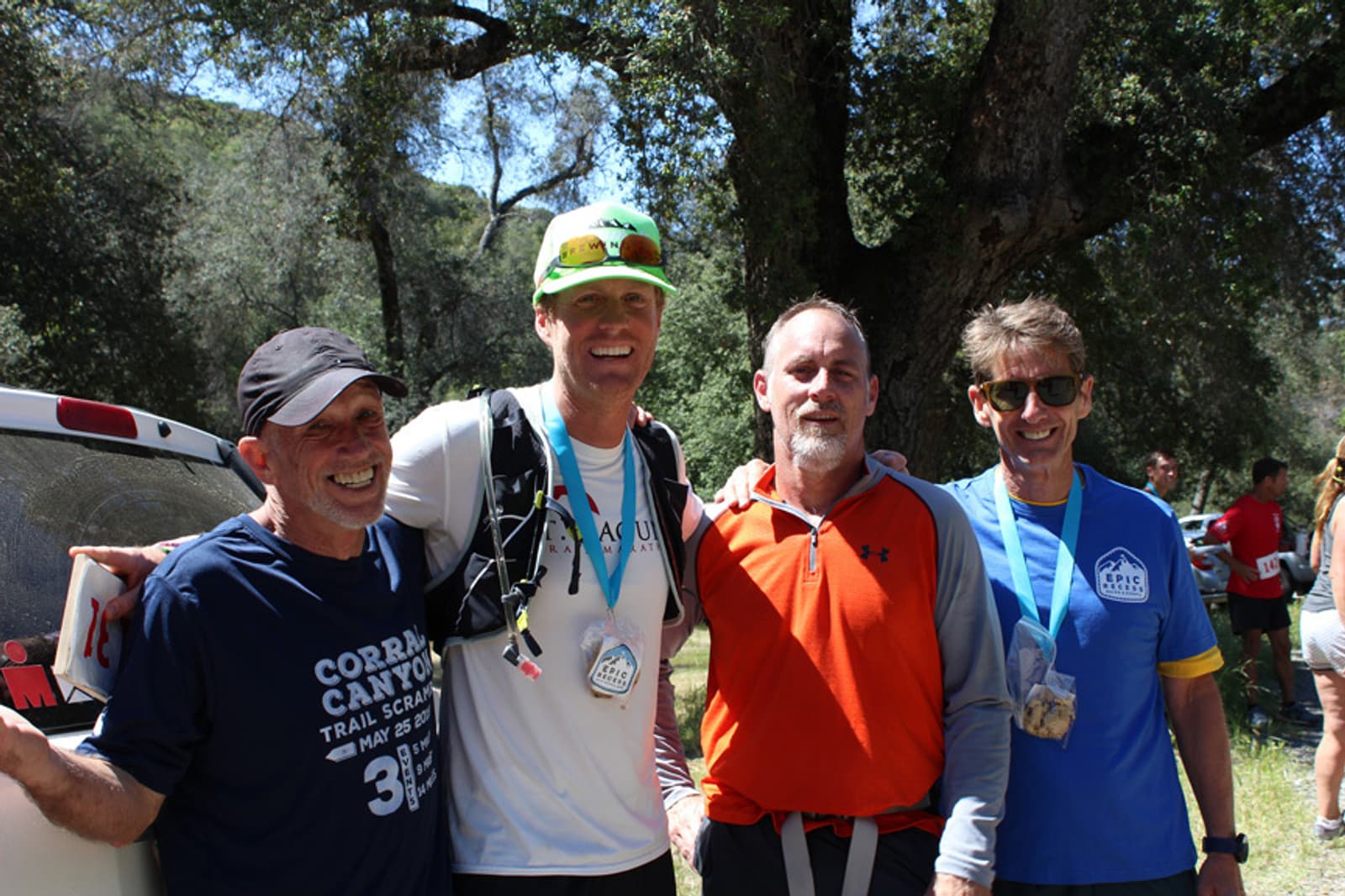 Four men are standing together outdoors, smiling at the camera. They are dressed in casual athletic clothing and wearing medals around their necks. Trees and sunlight are in the background.