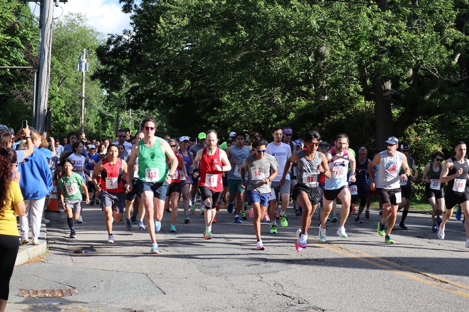 A group of runners participating in a race on a sunny day, surrounded by trees. They wear various athletic outfits and numbered bibs, running along a paved road. Spectators watch and take photos from the sidelines.