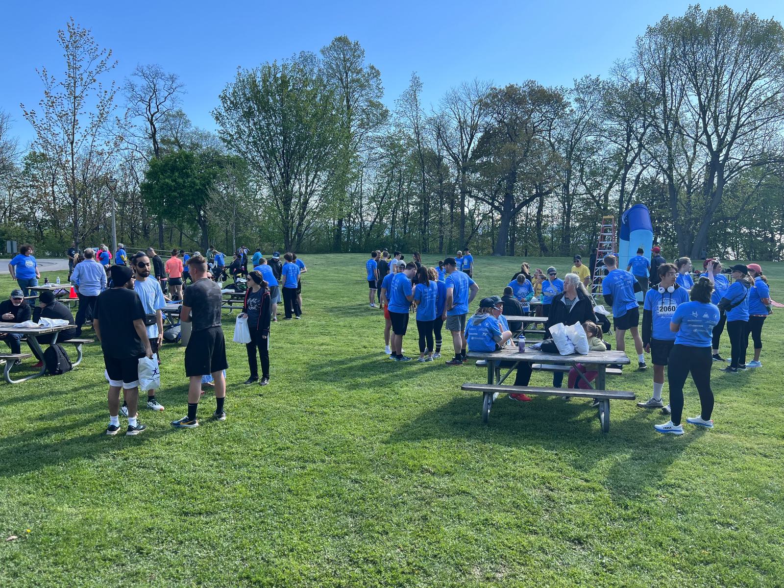A large group of people gathered in a park on a sunny day, with many wearing blue shirts. Some are sitting at picnic tables, while others are standing or walking on the grass. Trees and a bouncy structure are visible in the background.