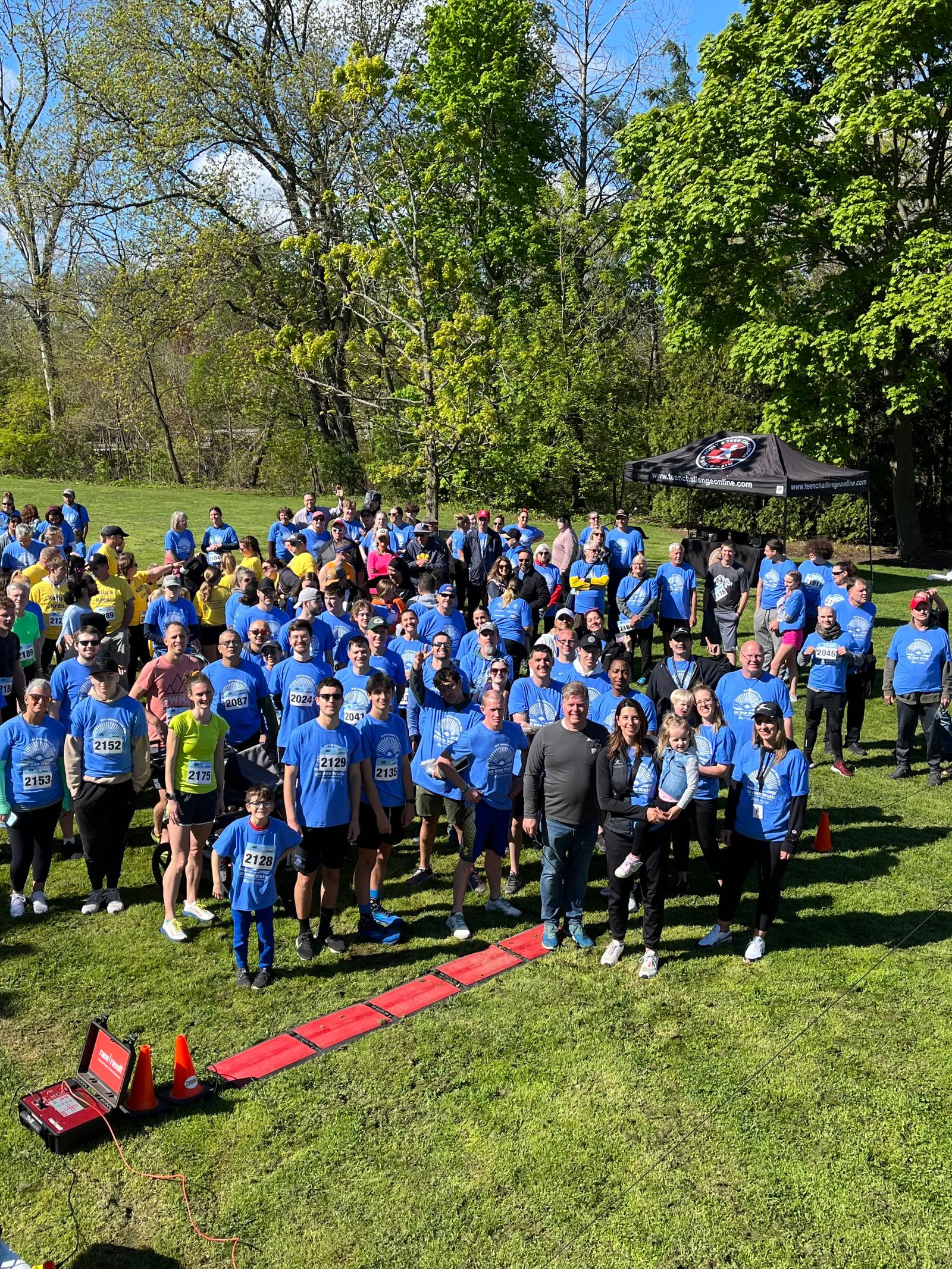 A large group of people wearing blue event shirts gather outdoors on grass, some with race bibs. A red timing mat is at the front, and trees and a tent are visible in the background under a clear sky.
