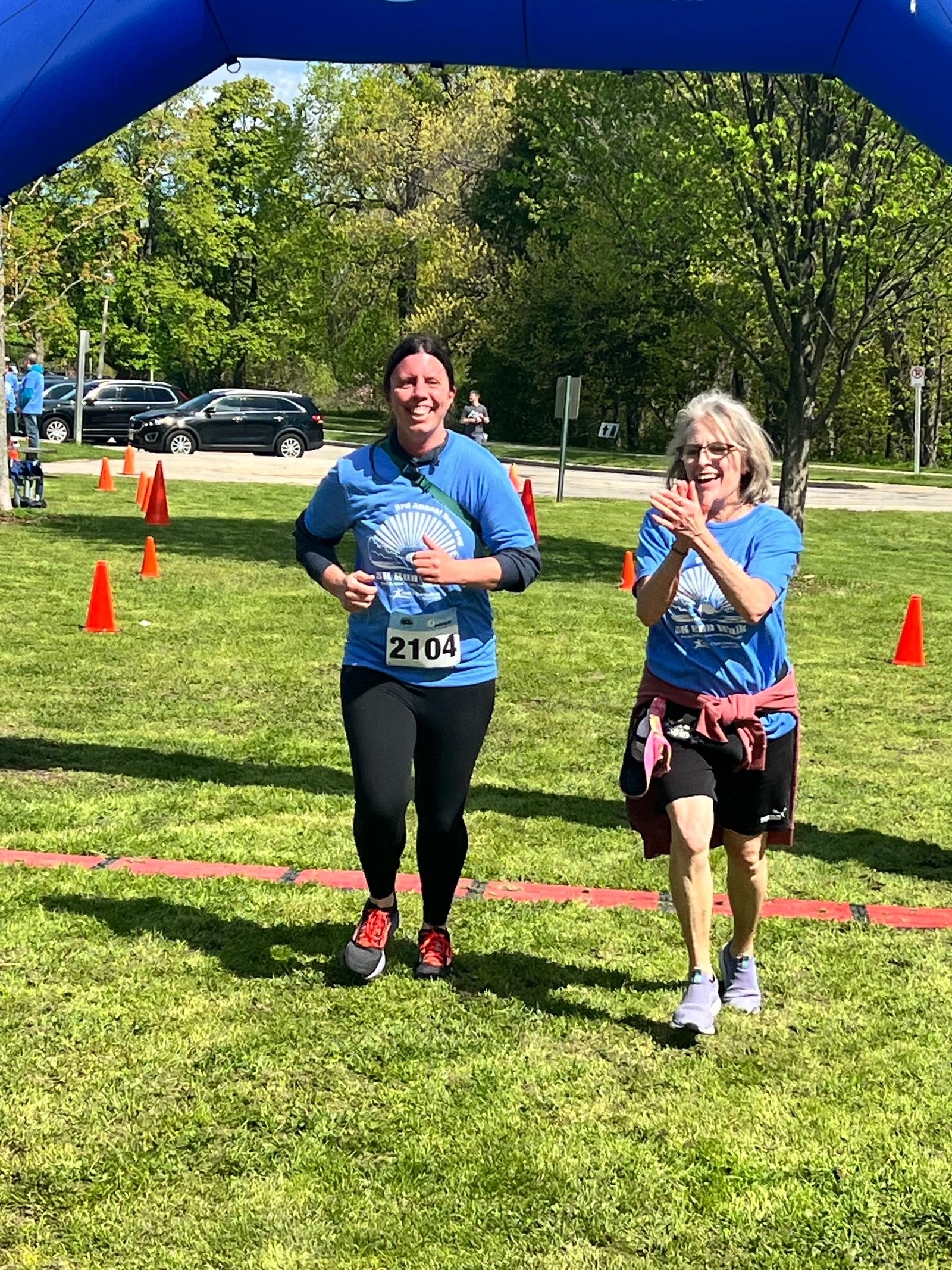 Two women in blue shirts, wearing race bibs, cross a finish line at an outdoor event. One woman claps while the other smiles. Orange cones line the grassy path. Trees and parked cars are visible in the background.
