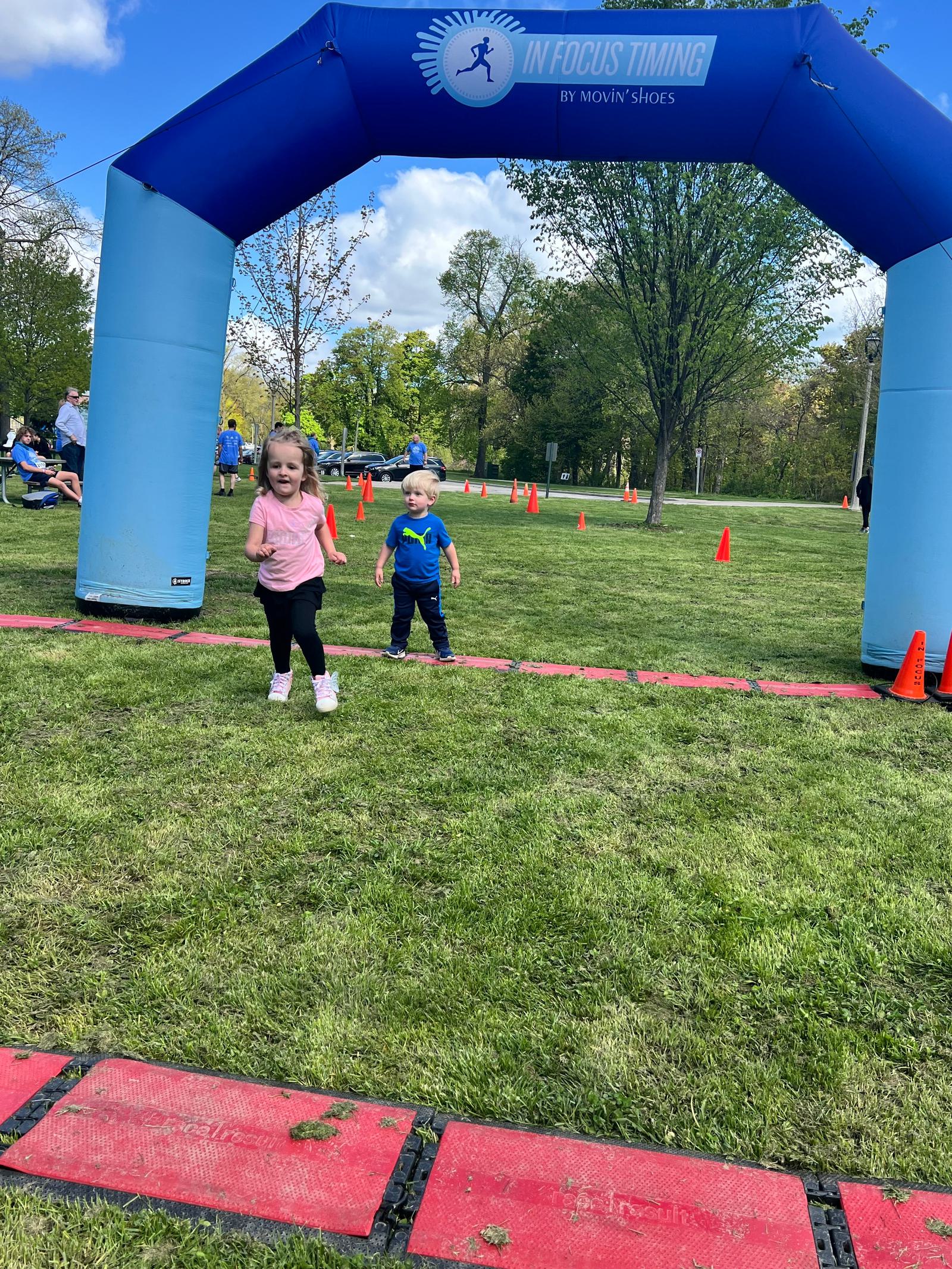 Two young children run toward a blue arch labeled "InFocus Timing" on a grassy field. Orange cones are set up around the area, and trees and scattered people are visible in the background. The sky is partly cloudy.