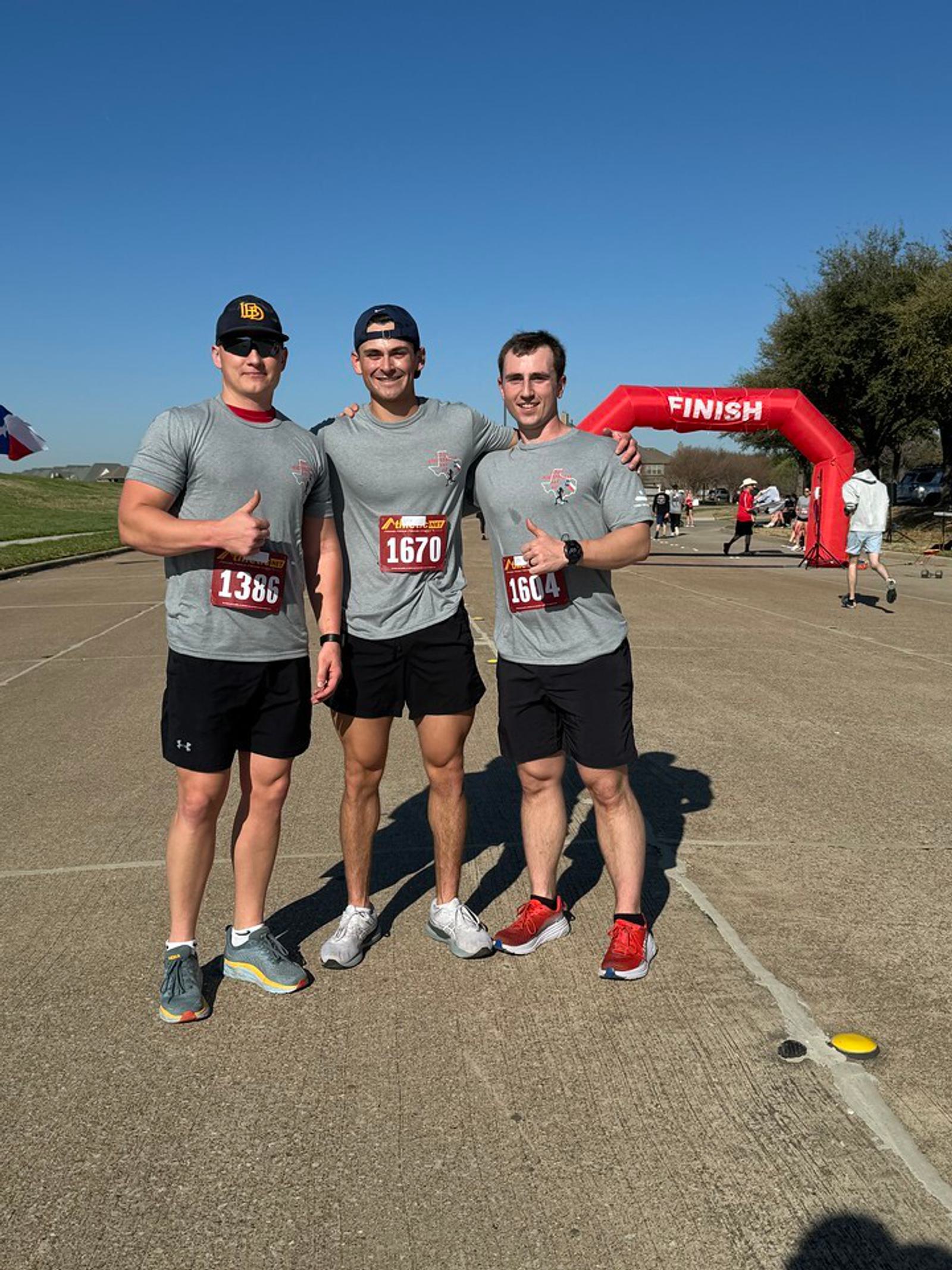 Three men in matching gray T-shirts pose with thumbs up near a red finish line arch at an outdoor race event. They wear running shoes and shorts, and two have race bibs. It's a clear, sunny day.