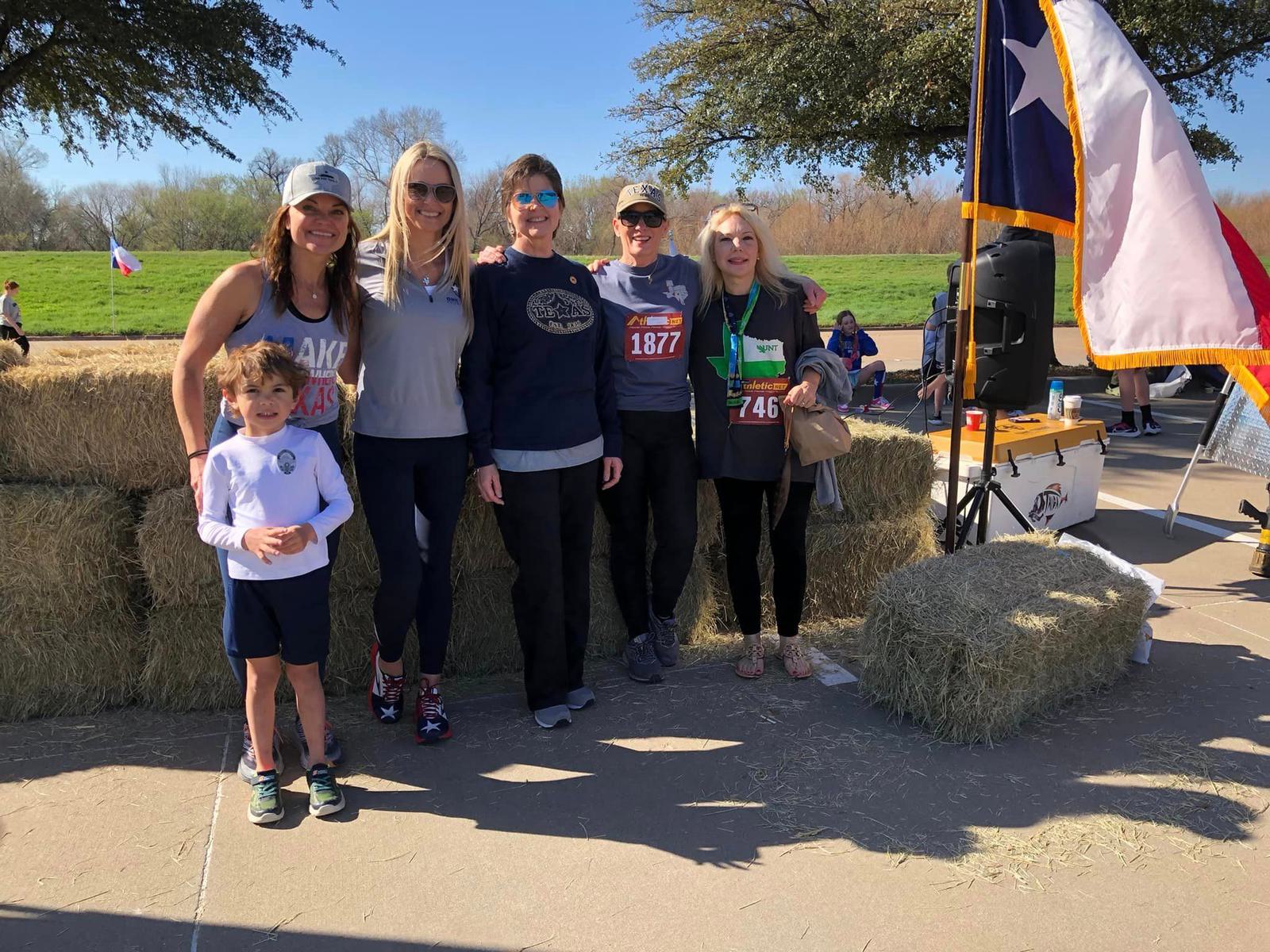 A group of five adults and one child pose for a photo near hay bales. Some are wearing race bibs. A Texas flag is displayed in the background, and the setting appears sunny and outdoorsy with trees and blue sky.