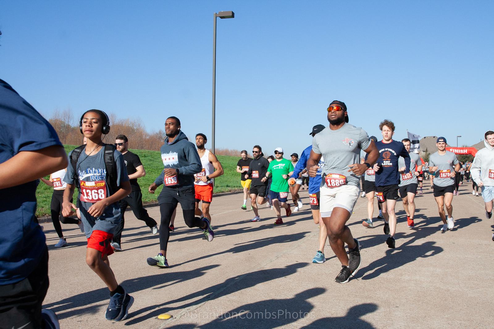 A group of runners in athletic clothing participates in a race on a sunny day. They are on a paved path with a clear blue sky in the background. Bib numbers are visible on their shirts.