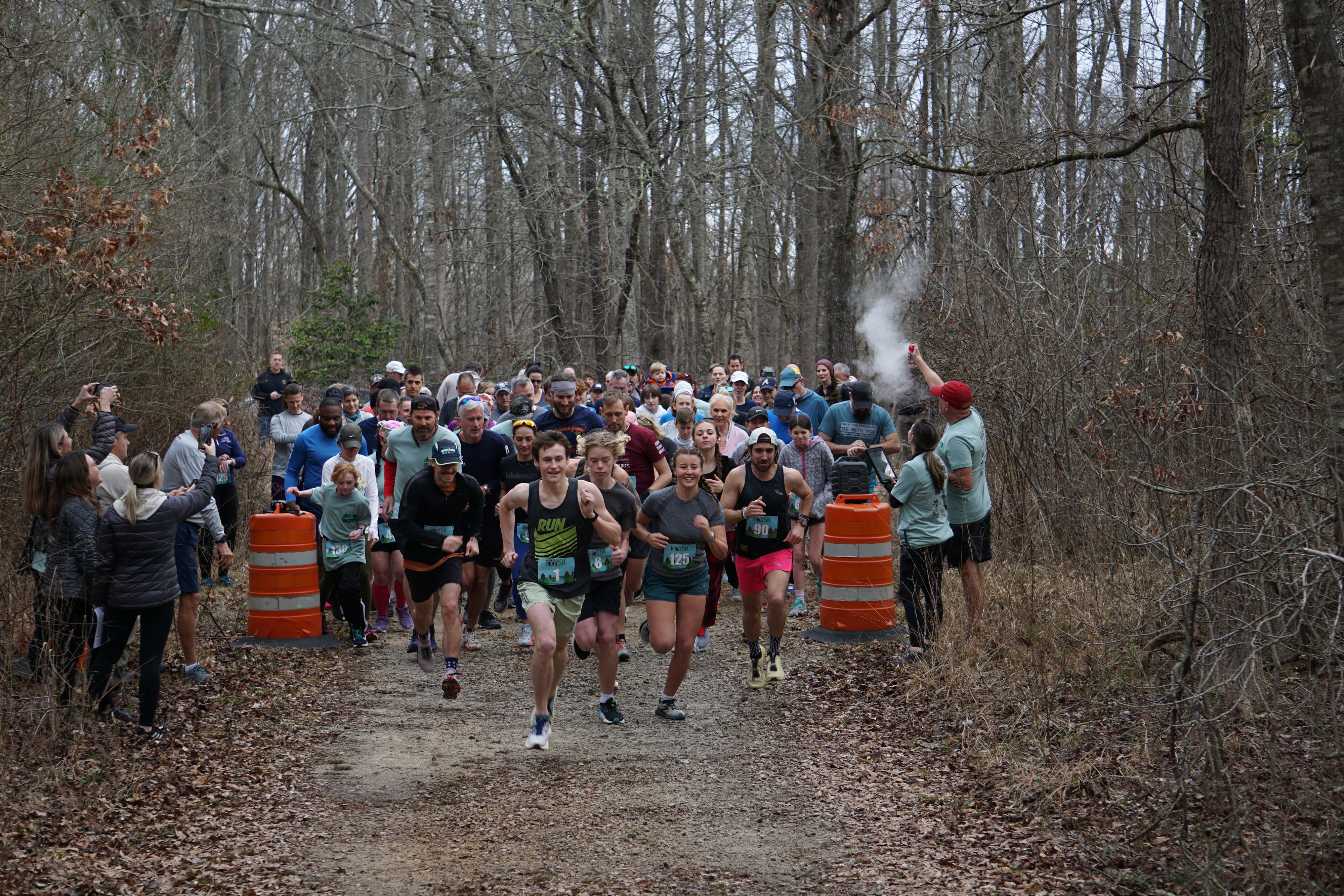 A large group of runners starts a race on a forest trail. A person on the right signals the start with smoke. Spectators line the edges of the path, and bare trees surround the scene on an overcast day.