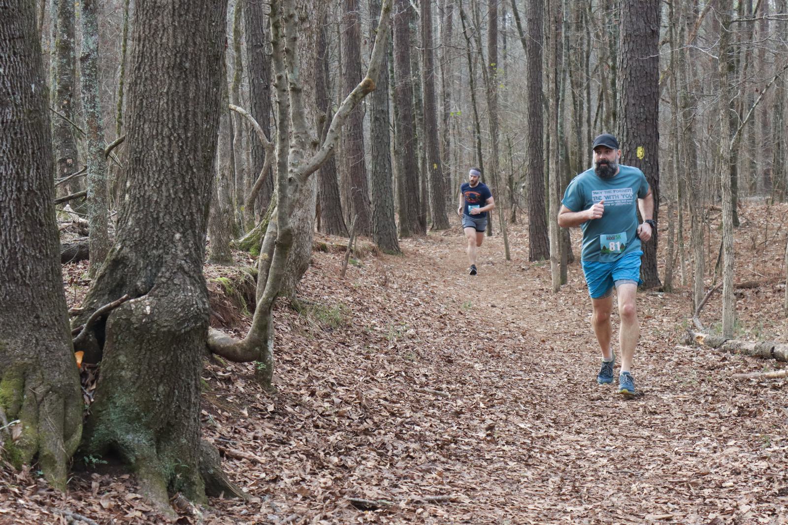 Two people are running on a leaf-covered trail through a forest of tall, bare trees. One runner is in the foreground wearing a blue outfit, while the other is further back. The scene is peaceful and natural.