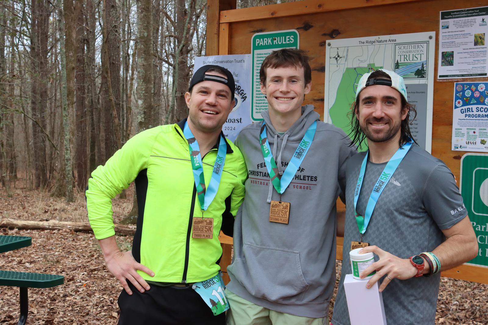 Three men wearing race medals stand smiling in front of a trail map. The man on the left wears a bright green jacket, the middle one a gray top, and the right one a gray t-shirt. They are in a wooded area, with leaves covering the ground.